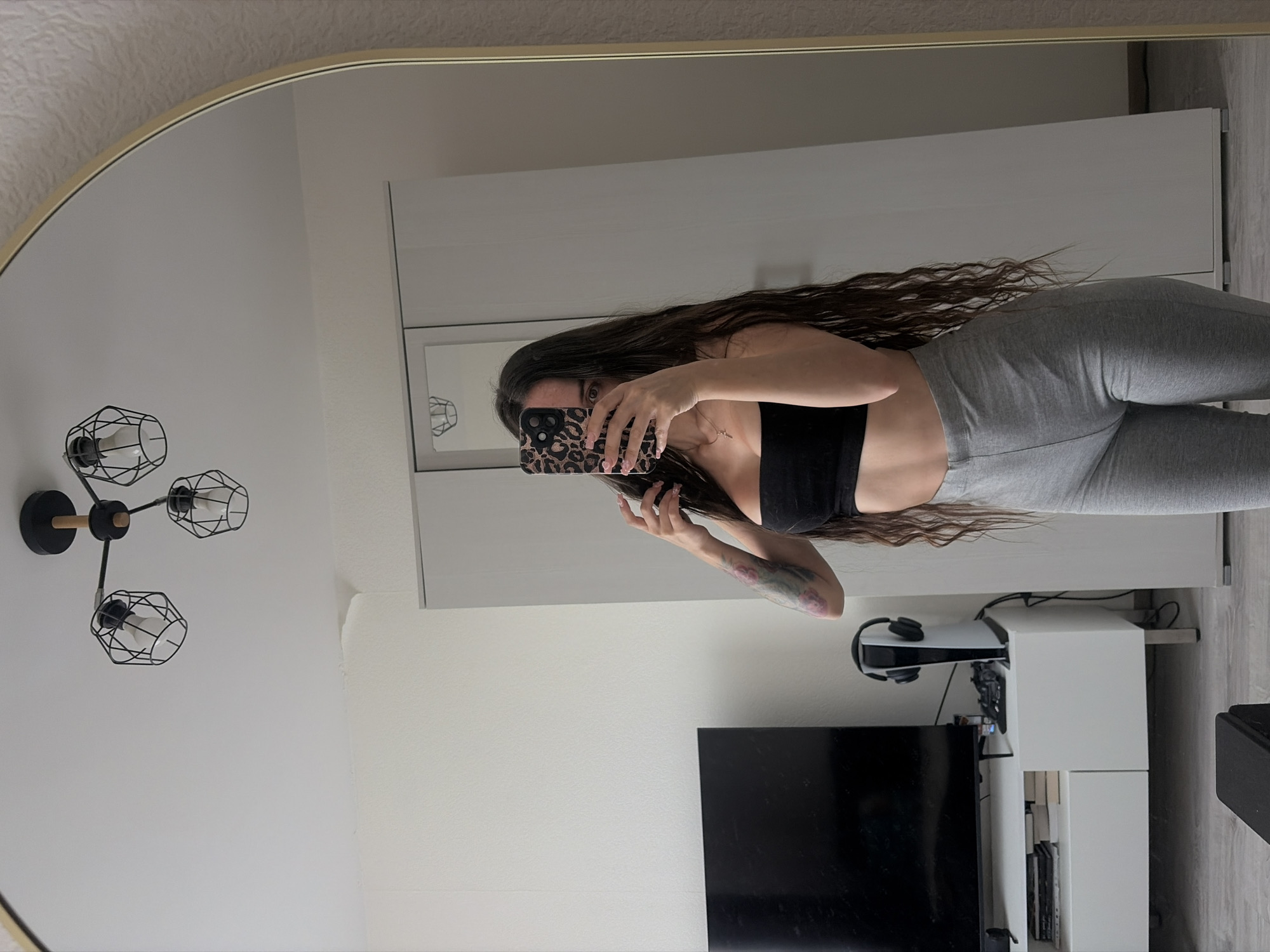 A young woman with long, dark, wavy hair is taking a mirror selfie in a room. She is wearing a black strapless top and grey leggings. She is holding a phone with a leopard print case in her right hand to obscure her face. Her left hand is touching her hair. A tattoo of pink roses is visible on her left arm. In the reflection of the mirror, a TV is visible on a white entertainment unit, along with a white PlayStation 5 console with headphones on top, and a black game controller. Behind the entertainment unit, a white closet is visible. A modern chandelier with black geometric shades hangs from the ceiling. The room appears to be a bedroom or living area with light-colored walls and a light-colored floor. The location is identified as Novosaratovka, Russia.