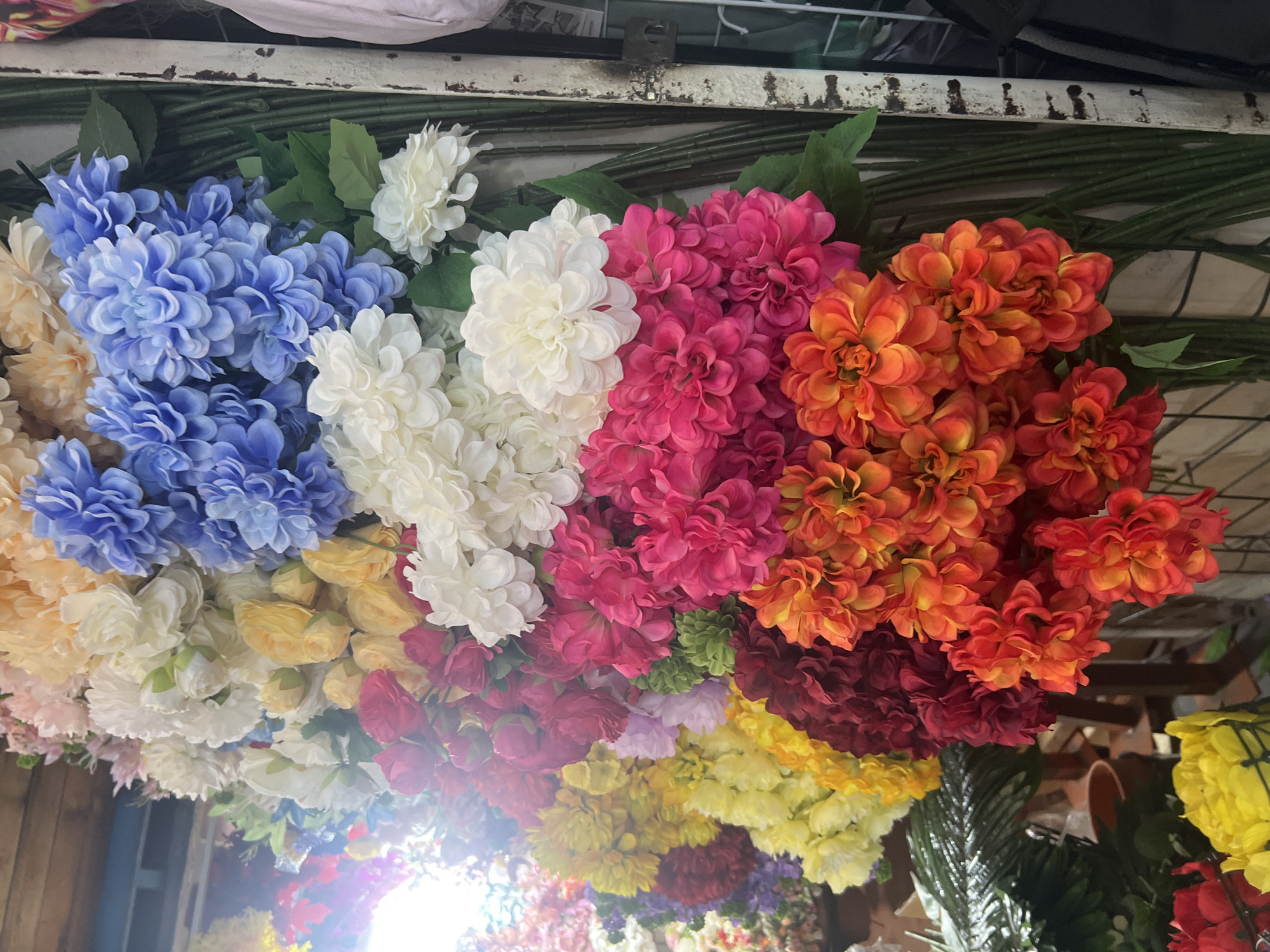 The image shows a close-up of a large display of artificial flowers. The flowers are arranged in bunches and are vibrant in color, including shades of blue, white, pink, orange, red, and yellow. They appear to be a variety of chrysanthemum or dahlia styles with full, layered petals. The flowers are densely packed and create a colorful, textured backdrop. In the background, there are hints of what appears to be a market stall or shop, with some greenery from artificial plants and what looks like a white pole or support structure with some weathering. The lighting suggests an indoor setting, possibly a market, with a bright light source coming from the left, creating some glare. There are no people visible in the image. The overall impression is of a vibrant and abundant display of decorative flowers, likely for sale.