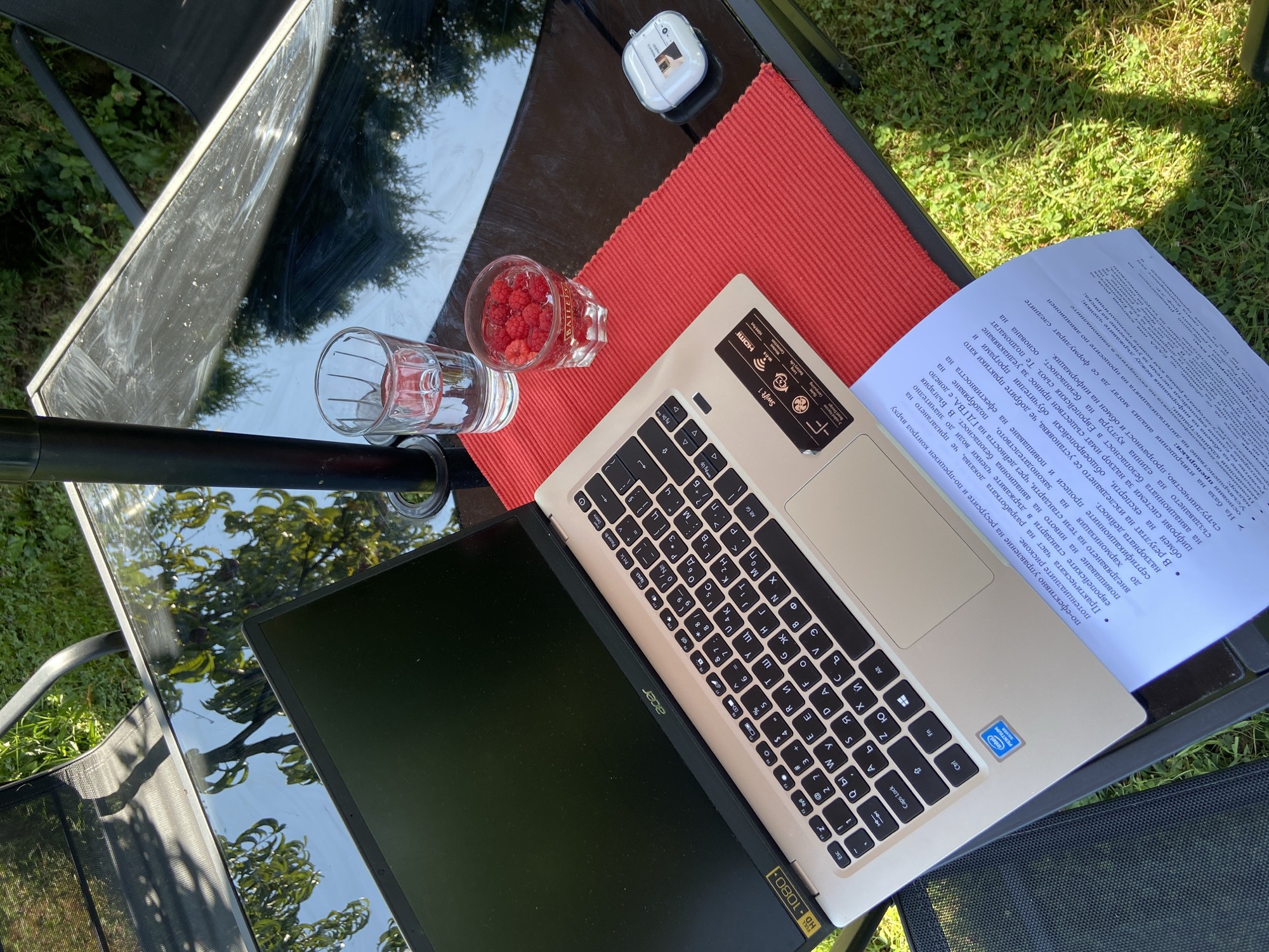 An overhead shot shows a laptop on a glass-topped outdoor table next to a glass of water and a glass of raspberries. The laptop is an Acer Swift 1. The table is set on a green, grassy lawn. A portion of a black outdoor chair is visible to the left. The scene appears to be outdoors in a residential setting, possibly a backyard. The location is specified as Mindia, Bulgaria. There are no people or animals present in the image. The focus is on the objects on the table and the natural environment surrounding it.