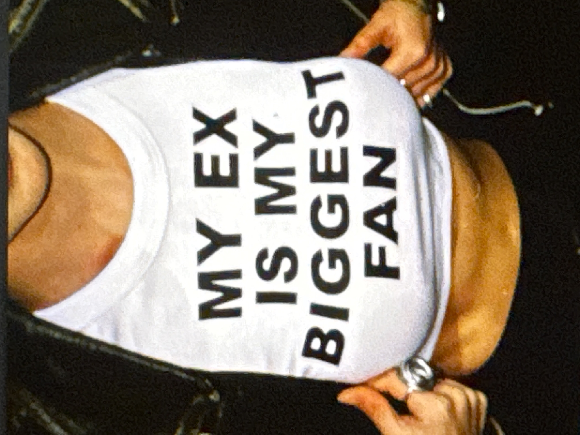 The image shows a close-up view of a person wearing a white tank top with bold black text. The text reads "MY EX IS MY BIGGEST FAN." The person's torso and a portion of their shoulder and chest are visible. They are wearing dark clothing underneath the tank top, possibly a jacket. Two hands are visible in the frame, one adjusting the tank top on the left side and another on the right, wearing rings. The lighting is somewhat dim, and the focus is on the text on the shirt. The image appears to be candid and taken from a high angle, looking down at the person. There are no clear indicators of time of day, weather, or specific location beyond what is implied by the provided location context of Kuta Selatan, Indonesia, which does not offer visual cues within the image itself. The overall mood is somewhat playful or defiant due to the statement on the shirt.