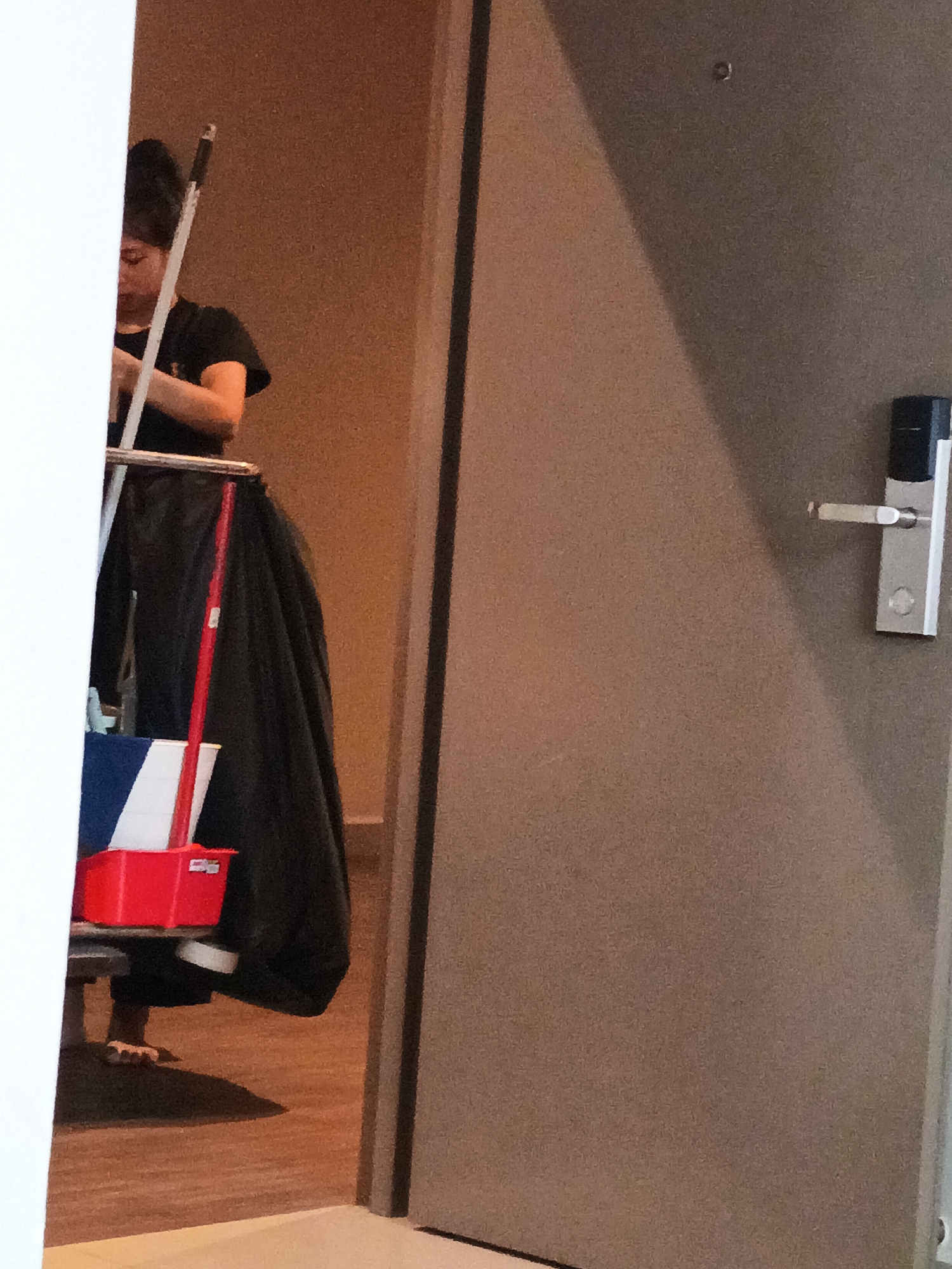 A person, likely a hotel staff member, is partially visible in a doorway on the left side of the frame. They are wearing a black t-shirt and have their hair tied up. They are pushing a cleaning cart that holds a red bucket with white and blue compartments. A black trash bag is also attached to the cart. The person is barefoot, indicating they might be in the process of cleaning a room or are on break. The background shows a wall and a door with a silver handle on the right side. The lighting suggests an indoor setting, possibly a hotel corridor in Kuala Lumpur, Malaysia. The overall scene depicts a mundane moment of daily work.