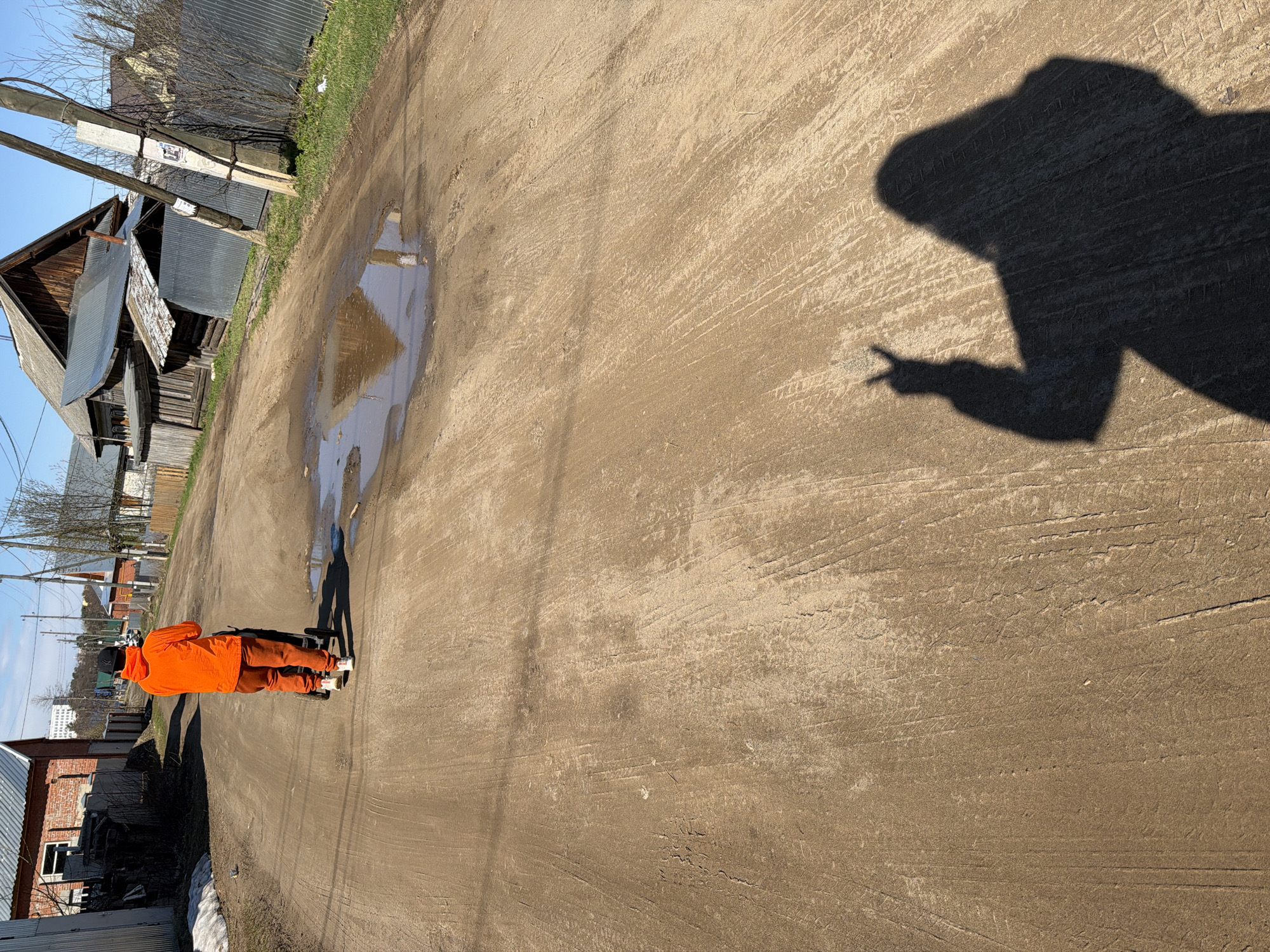 The photograph appears to be taken on a dirt road in a rural or semi-rural area, possibly on the outskirts of a town or village. Based on the architecture of the wooden houses and the overall aesthetic, it could potentially be in Chaikovsky, Russia.
The subject, a person wearing an orange hooded jacket and dark trousers, is standing on the road. Their shadow is cast on the ground in front of them, suggesting the sun is behind them. The shadow's hand is making a "peace" sign with two fingers.
The road itself is uneven, with visible tire tracks and a puddle of water reflecting the sky. The surrounding area features wooden houses with corrugated metal roofing.
In the background, there's a wooden house to the left of the frame with what appears to be a damaged roof. There's also a telephone pole. On the right side, more houses are visible along with a metallic fence. The sky is blue, suggesting a clear day.