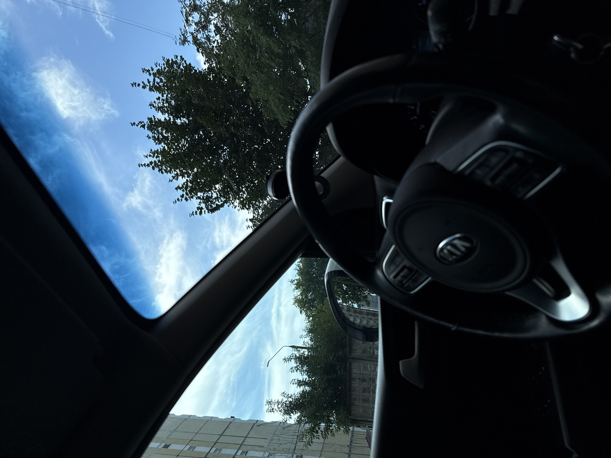 An interior view from a vehicle shows a steering wheel with the Kia logo prominently displayed. The dashboard is visible, along with a car door handle on the left. Through the windshield and side windows, a cloudy blue sky is visible. Outside the car, there is a light-colored building on the left, a concrete fence, and lush green trees. A lamppost stands behind the fence. The scene appears to be captured in the city of Korolyov, Russia.