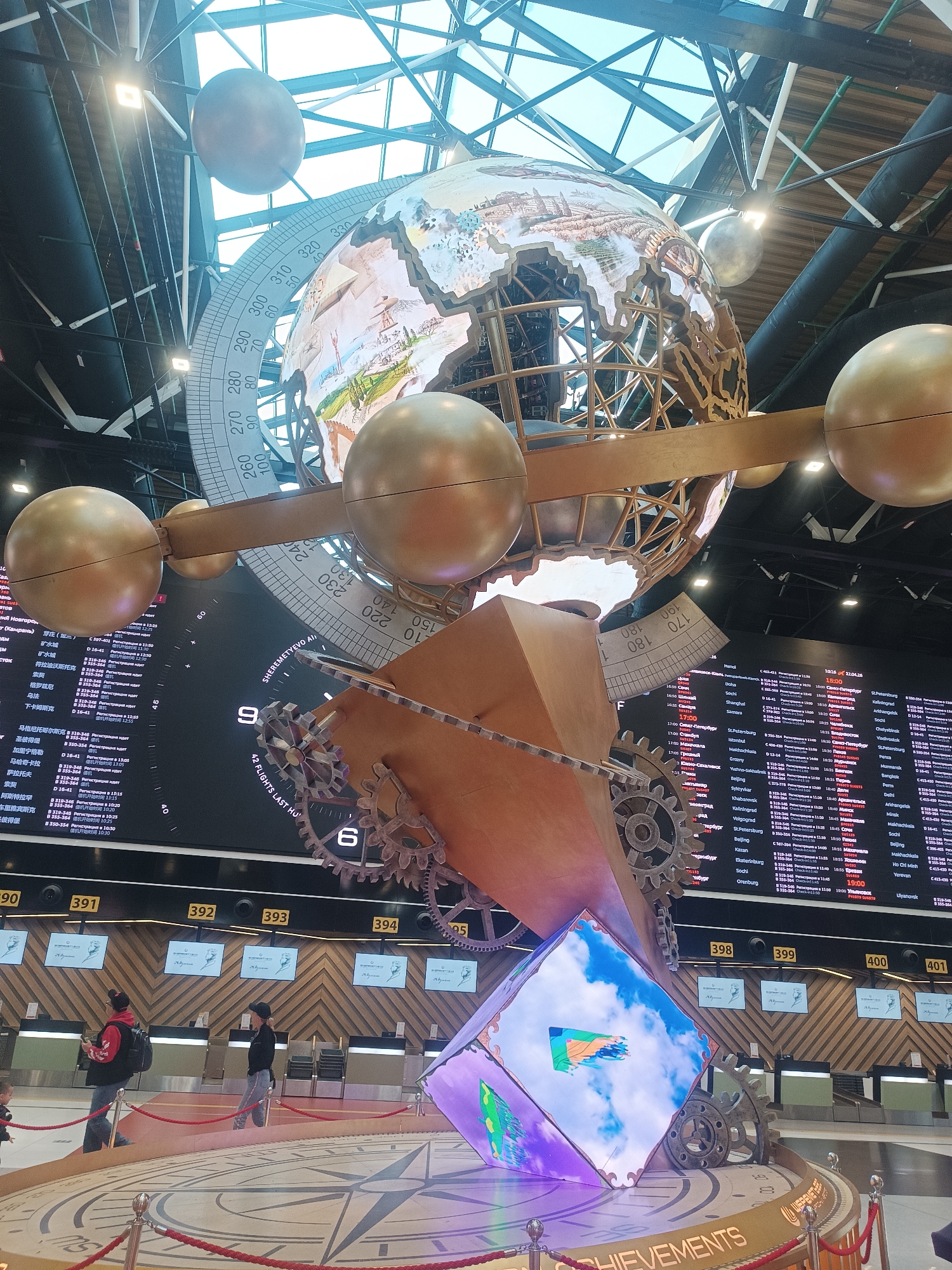 The image captures a grand, intricate art installation within what appears to be a modern airport terminal. At the center, a large, ornate globe with a detailed map (partially depicting landscapes) is suspended, encircled by a massive protractor-like dial marked with numbers from 0 to 330. Several smaller, smooth golden spheres orbit the globe, connected by a brass-colored structure, reminiscent of an orrery. Below this celestial model, a complex mechanical structure featuring large gears, a pointer, and a rectangular section with angular cutouts dominates the mid-ground.
At the base of the installation, a multi-faceted LED screen displays vibrant, colorful imagery, including what looks like a blue sky with clouds and abstract patterns. This screen rests on a circular base adorned with a compass rose design and the word "ACHIEVEMENTS." The entire structure is roped off with red velvet ropes.
In the background, large digital display boards show flight information, including destinations, flight numbers, times, and statuses in both Cyrillic and Chinese characters. Visible destinations include St. Petersburg, Kaliningrad, Beijing, Istanbul, and Ho Chi Minh. One board clearly displays "SHEREMETYEVO" and "10:16 22.04.26," indicating the airport and a date. Another segment reads "42 FLIGHTS LAST HO." Below these screens, a line of check-in counters, numbered from 390 to 401, is visible. Several people, including adults and a child, are seen near these counters, suggesting typical airport activity such as waiting or checking in.
Overhead, a bright glass ceiling with a visible metal framework allows natural light to illuminate the space, suggesting it is daytime. The overall scene conveys a sense of grandeur and technological sophistication, characteristic of a major international airport like Sheremetyevo in Khimki, Russia.