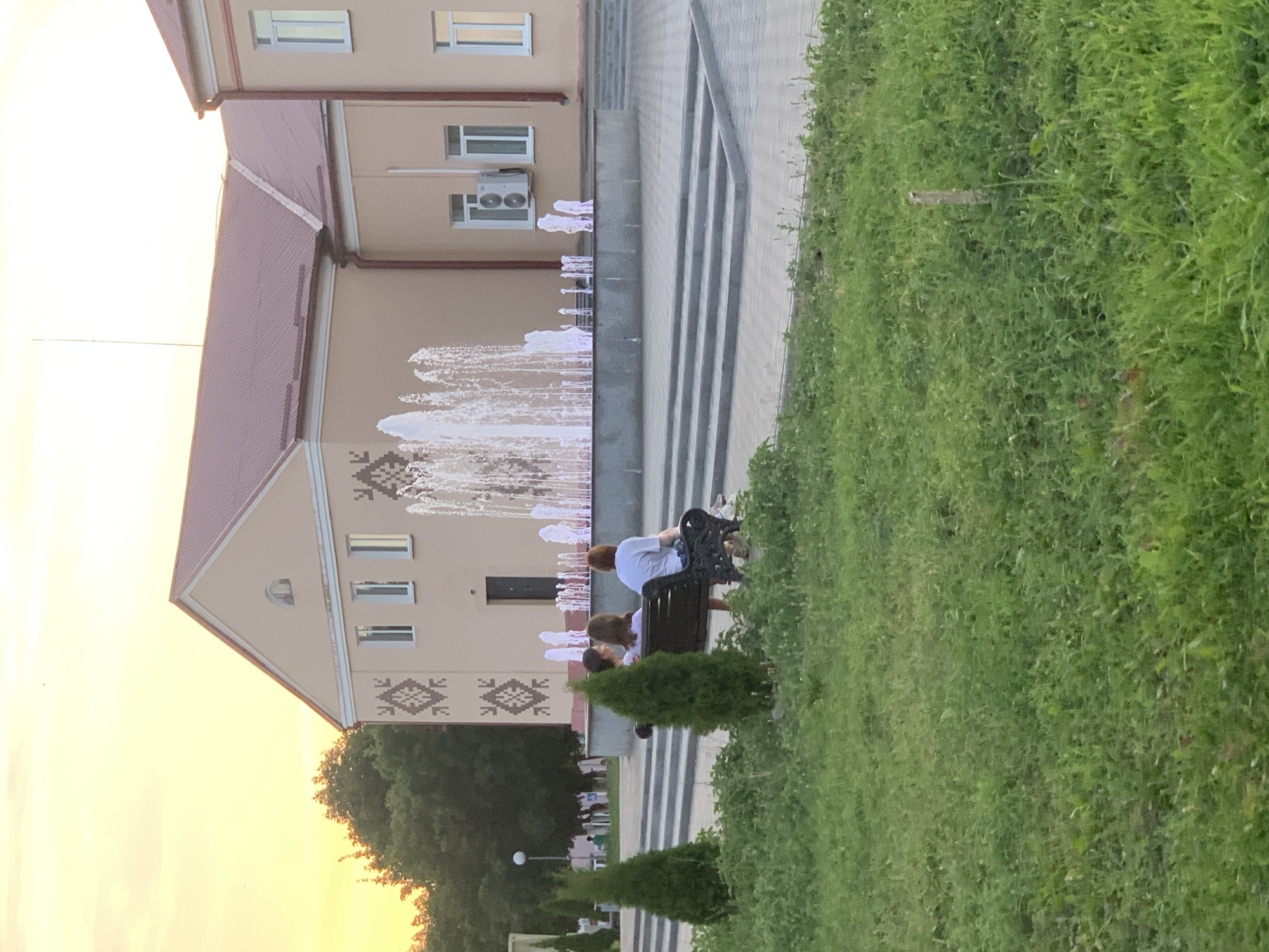 The image depicts a public space in Kapyĺ, Belarus, featuring a building with light beige walls and a red roof. The building has several windows, and an air conditioning unit is visible on one section of the facade. In front of the building, a fountain sprays water, illuminated with pink and white lights. To the left of the fountain, a group of people are seated on a dark bench, facing away from the camera. Three conical evergreen trees are planted in front of the bench. The foreground is a grassy area, and in the background, there are more trees and a lamppost. The sky is a warm, pale yellow, suggesting it might be sunset or sunrise. The area around the fountain and building is paved with grey tiles and features stepped seating areas.