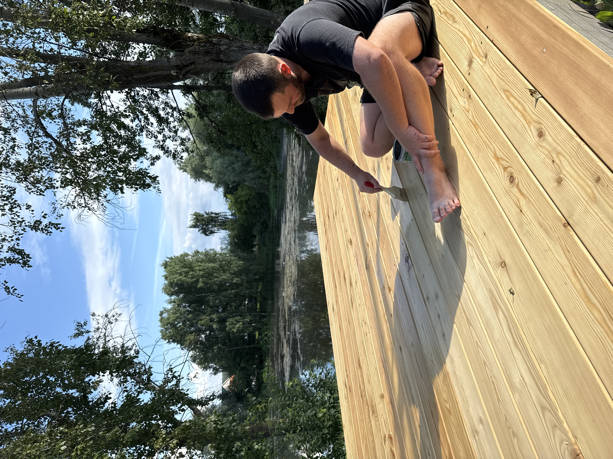 A man with a beard, wearing a black t-shirt and dark shorts, is sitting on a newly constructed wooden deck, painting it with a brush. He is right-handed and is applying a transparent stain to the wooden planks. A can of stain is next to him on the deck. His bare feet are visible. The deck appears to be near a body of water, possibly a river or lake, with trees and vegetation on the opposite bank. The sky is blue with scattered clouds. The location is specified as Chaikovsky, Russia.