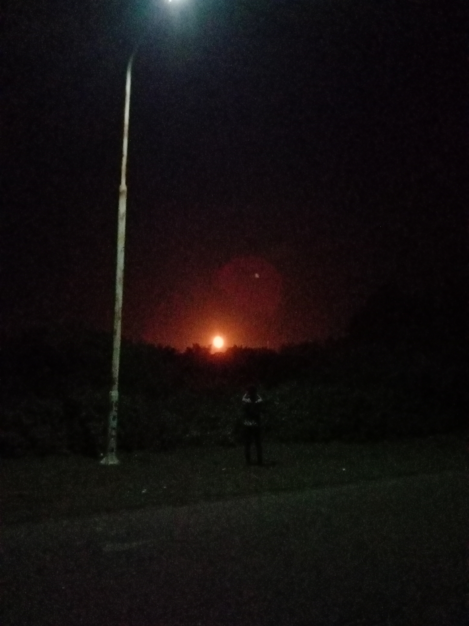 The image shows a nighttime scene in an outdoor environment, possibly in Lhokseumawe, Indonesia. A tall light post stands on the left side of the frame, illuminated at the top. In the distance, there is a bright orange glow, possibly from a fire or industrial activity, situated on the horizon behind a line of trees or vegetation. A silhouetted figure, possibly a person, is standing in a grassy area, seemingly facing the orange glow. The foreground consists of a dark paved surface or road. The overall atmosphere is dark, with the artificial light and the distant glow providing the only sources of illumination.