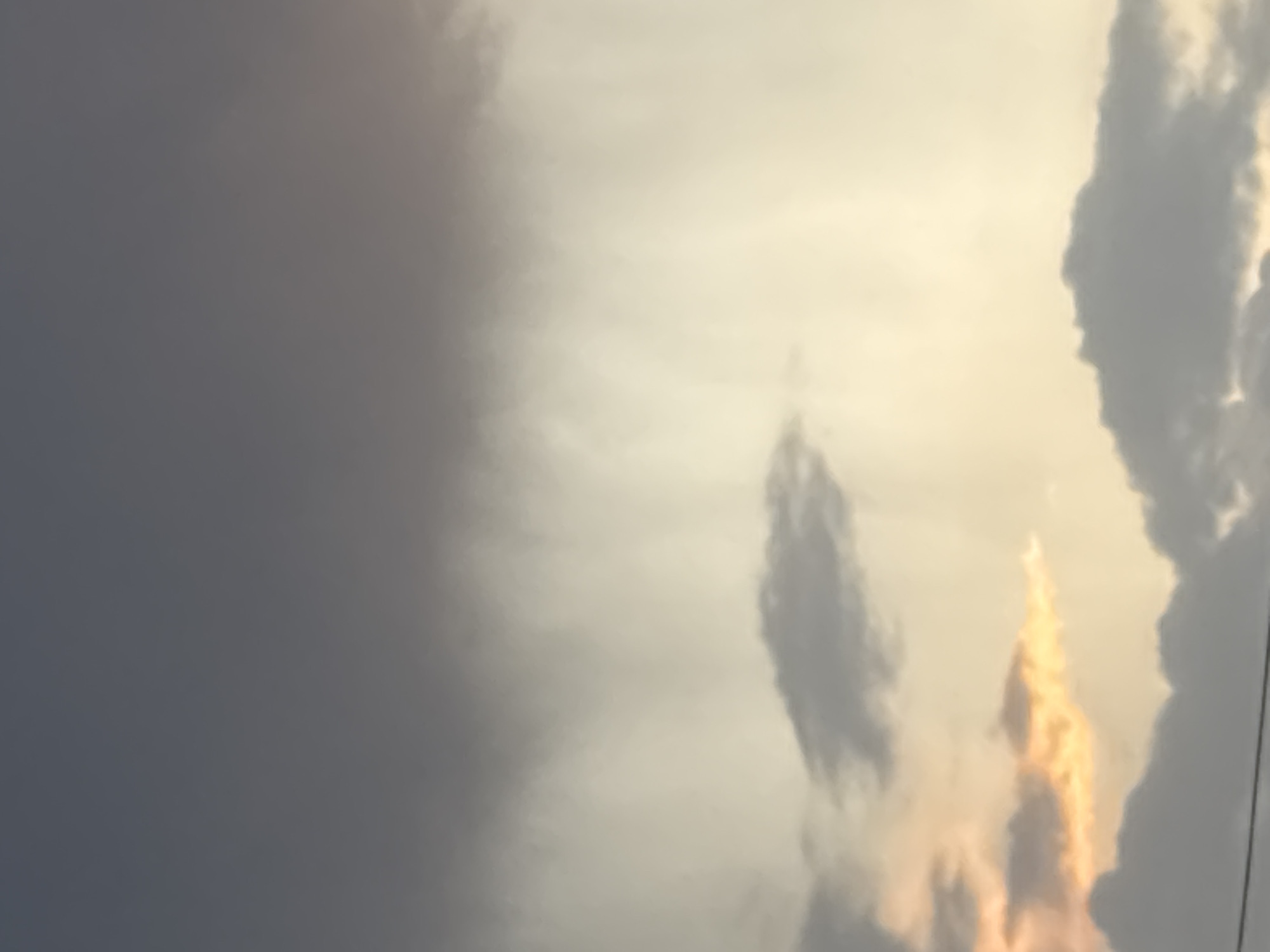 The image shows a sky scene. The sky transitions from dark gray clouds on the left to brighter, yellowish-white clouds on the right. There's a vertical formation of darker clouds in the center, and near the right edge, a patch of clouds is illuminated with an orange-yellow glow. Thin dark lines appear near the rightmost edge. The location, Pchelnik, Bulgaria, is not visibly represented in the image.
