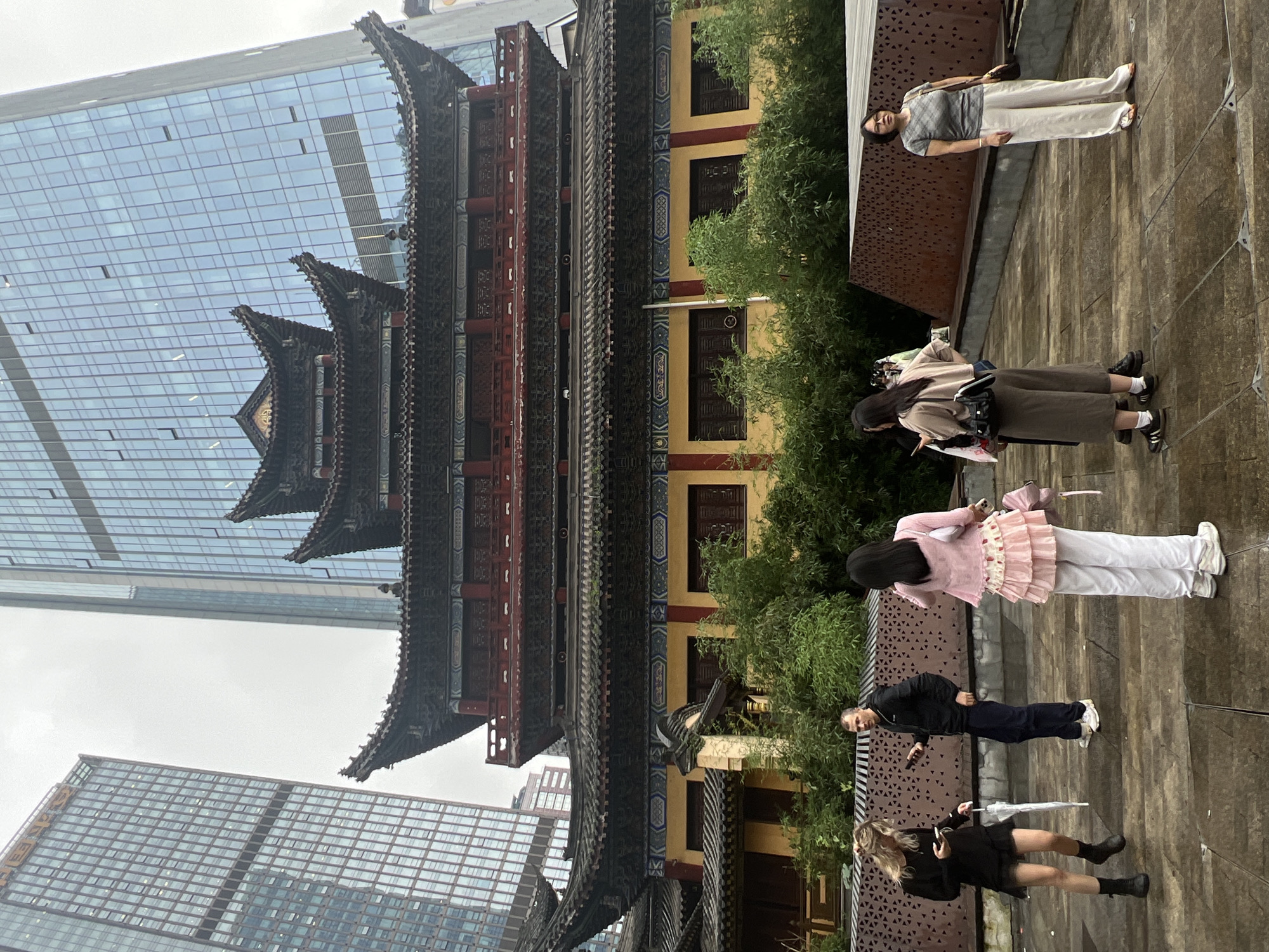 The image depicts a serene outdoor scene, likely in Yuzhong District, China, blending traditional architecture with modern elements. In the foreground, several individuals are captured from a slightly elevated perspective. One woman, dressed in a white top and light-colored pants, stands on a paved area, holding a dark bag. Further down, two young women are positioned. One, in a pink cardigan and a frilly white skirt, holds an umbrella. The other, in a black dress and boots, also holds an umbrella. A man in a black jacket and dark pants is between them, looking towards the camera and holding a phone.
Behind the people, a striking traditional Chinese building with ornate dark tiled roofs and red and yellow accents stands prominently. This architectural marvel is juxtaposed against a backdrop of contemporary glass skyscrapers, suggesting a location where old and new coexist. Lush green trees partially obscure the traditional building, adding a natural element to the scene.
The lighting suggests it might be a cloudy day, possibly in the late afternoon or early morning, as there are no strong shadows. The overall atmosphere appears calm and contemplative. There is no visible text in the image.