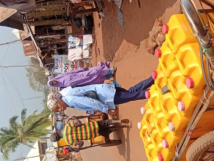 A street scene shows a cart loaded with numerous yellow jerrycans, each capped with red. Beside it, two women in headscarves, one light blue and one lavender, stand on the street. A man in a striped shirt is pulling a cart with small wheels. Businesses and merchandise are visible on the side of the street.