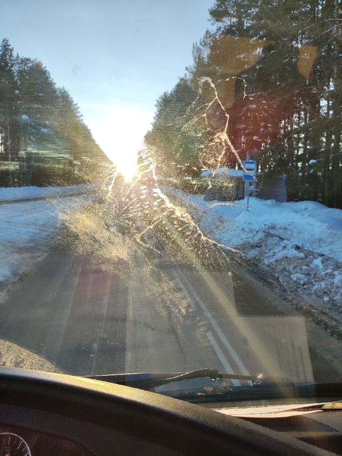 The view from inside a vehicle shows a road ahead through a dirty windshield. The sun shines brightly at the end of the road, between lines of trees. Snow sits on the side of the road, accumulating near a bus stop. The sun's glare is reflected and distorted on the windshield, making details difficult to discern clearly. Windshield wipers are visible in the foreground.