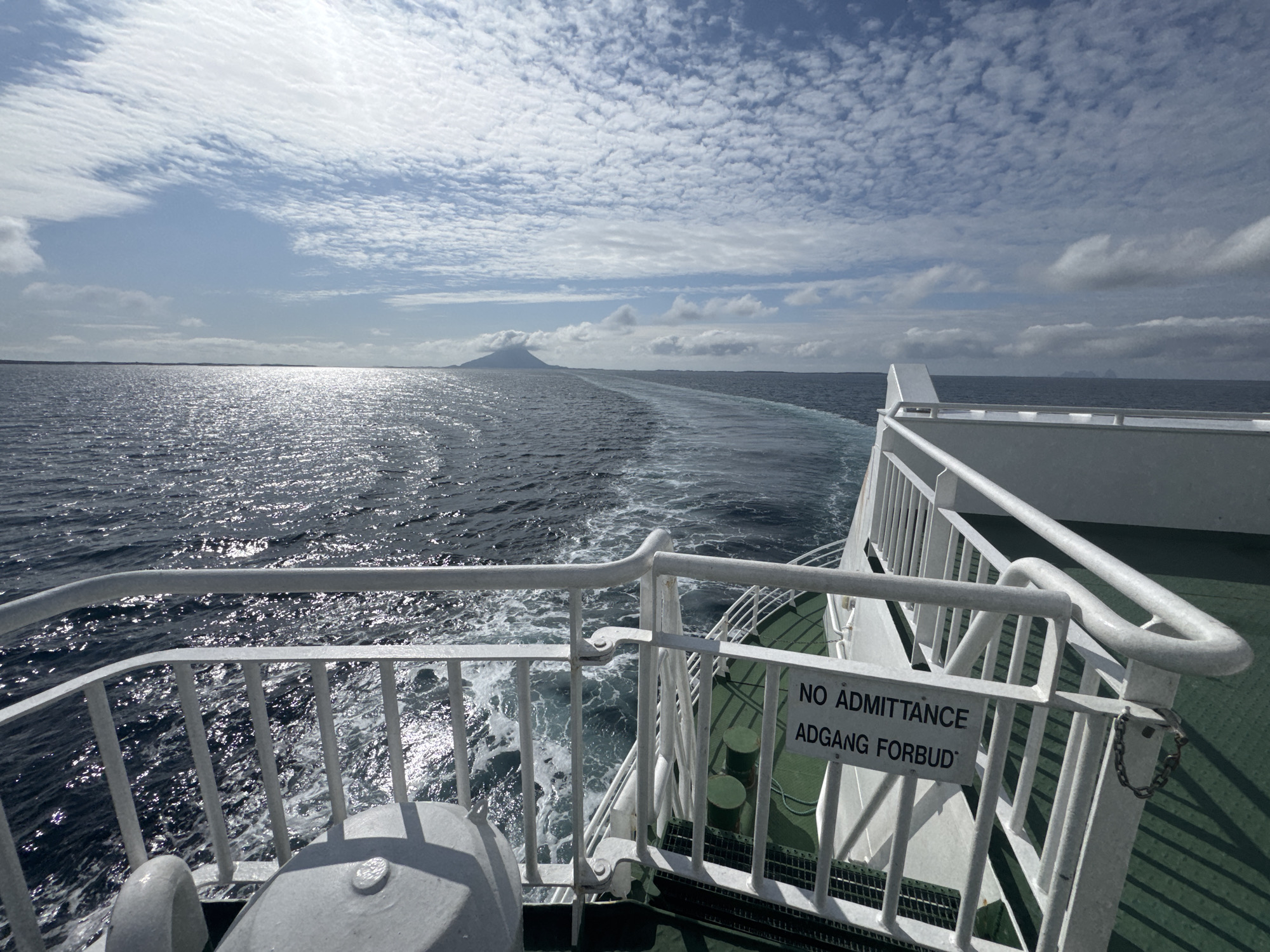 The image depicts a view from the stern of a ferry sailing on the ocean. The water is choppy with a visible wake trailing behind the vessel. In the distance, a conical mountain rises from the sea, possibly indicating an island or coastal feature. The sky is filled with scattered clouds, and the sun reflects brightly on the water's surface.
On the ferry's deck, a white railing lines the edge, with a sign attached that reads "NO ADMITTANCE ADGANG FORBUD". The deck itself is a dark green color. The overall scene is one of travel on the water under a partly cloudy sky. The location is identified as being near Sleneset, Norway.