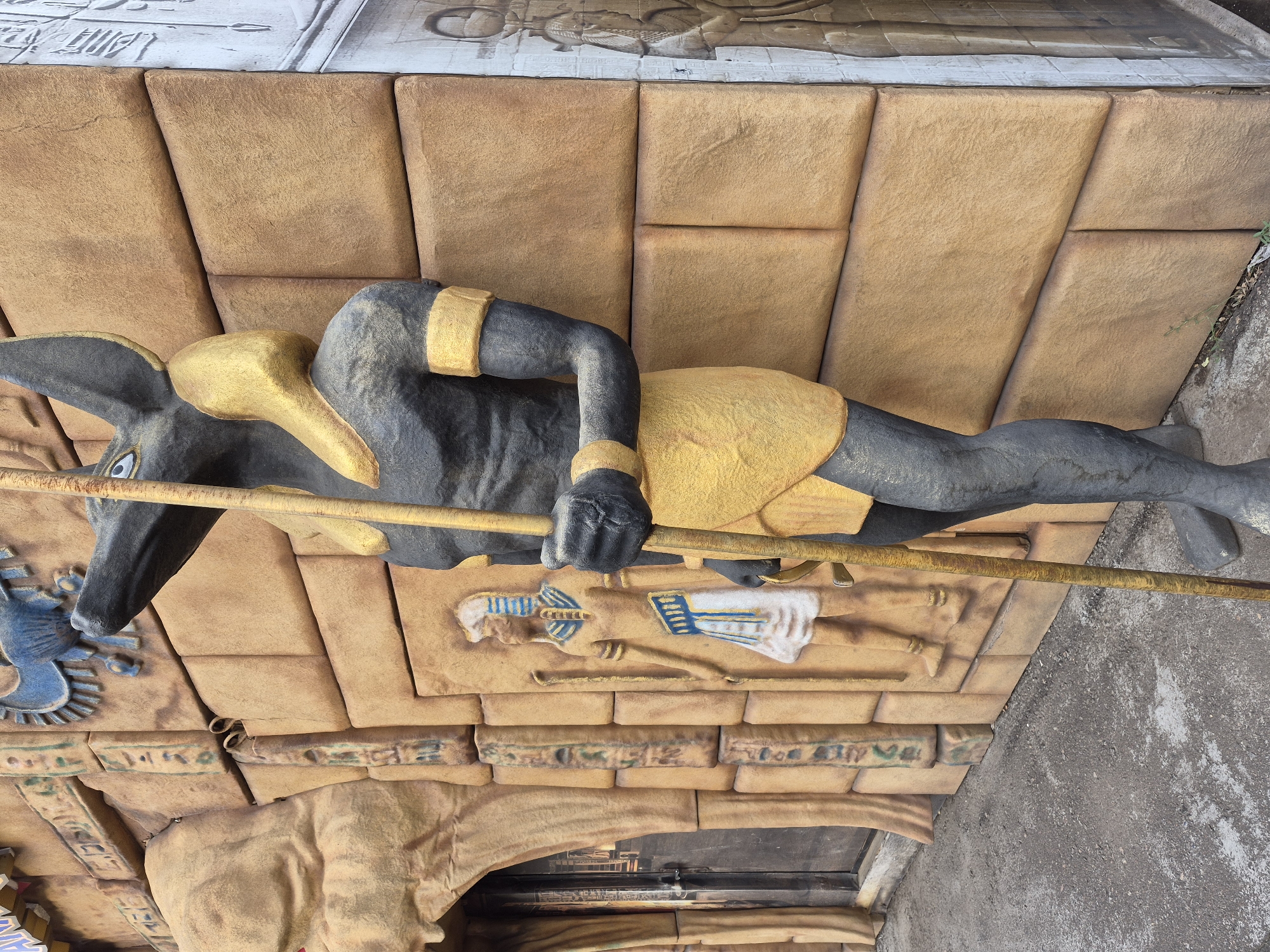An outdoor, medium-shot, eye-level, full-body statue of the Egyptian god Anubis stands next to a building with Egyptian-themed architecture. The statue, made of dark gray and gold material, depicts Anubis with a jackal's head, golden collar, and a yellow kilt, holding a wooden staff. The building's facade is made of tan, textured blocks that resemble ancient stone, with hieroglyphic carvings and a relief of a pharaoh. To the left of the statue is an entrance with a dark door and more Egyptian carvings. The ground in front of the building is paved with gray concrete. The lighting suggests it is daytime.