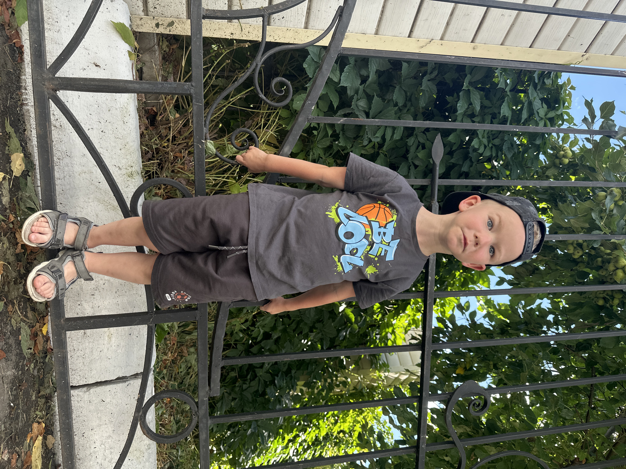 A young boy, approximately 4-5 years old, stands outdoors near a black wrought-iron fence. He is wearing a dark gray t-shirt with a graphic design that includes a basketball and the text "BE COOL," dark gray shorts, a black baseball cap, and gray sandals. The boy is holding a small, dark object in his right hand. Behind him, lush green foliage and a blue sky are visible. The fence has decorative scrollwork and appears to be in front of a building, as suggested by the presence of a white, horizontally sided wall to the left of the image. The ground in front of the fence is covered with dirt and fallen leaves. The setting appears to be a residential area in Даниловка (алтайский), Russia, likely a yard or garden. The lighting suggests it is daytime.