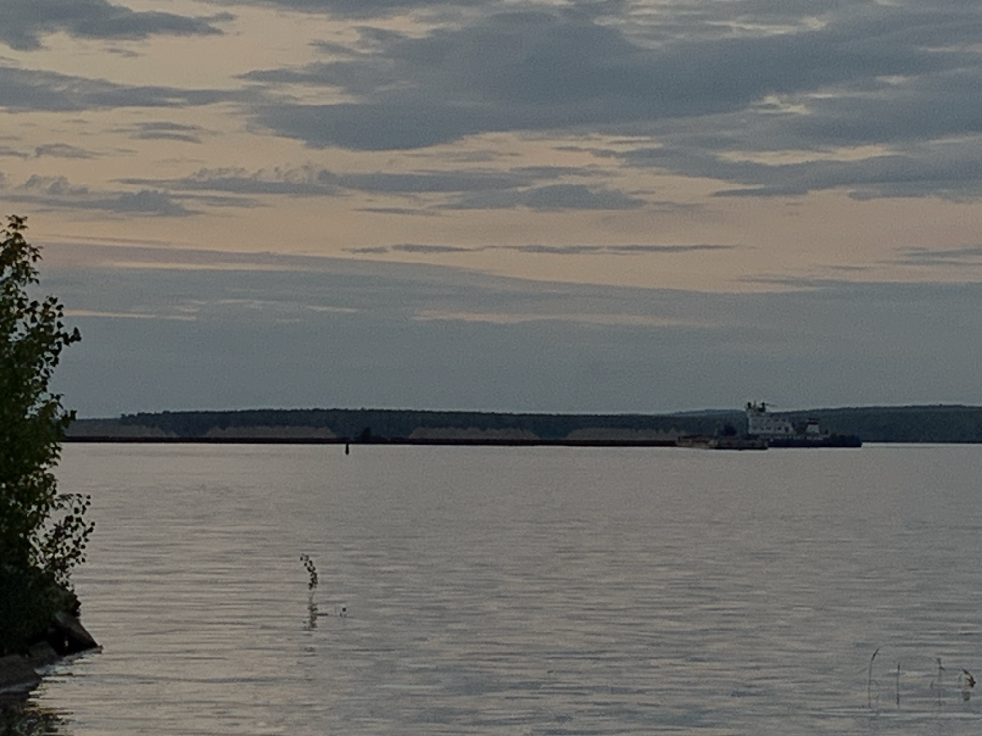 The image depicts a body of water, likely a river or reservoir, under a cloudy sky. On the far side of the water, a long barge laden with material is visible, accompanied by a tugboat at its stern. A wooded shoreline forms the background. In the foreground on the left, the edge of a bank with vegetation is present. The water exhibits subtle ripples. This scene is located on the Kama River in the city of Chaikovsky, Russia.