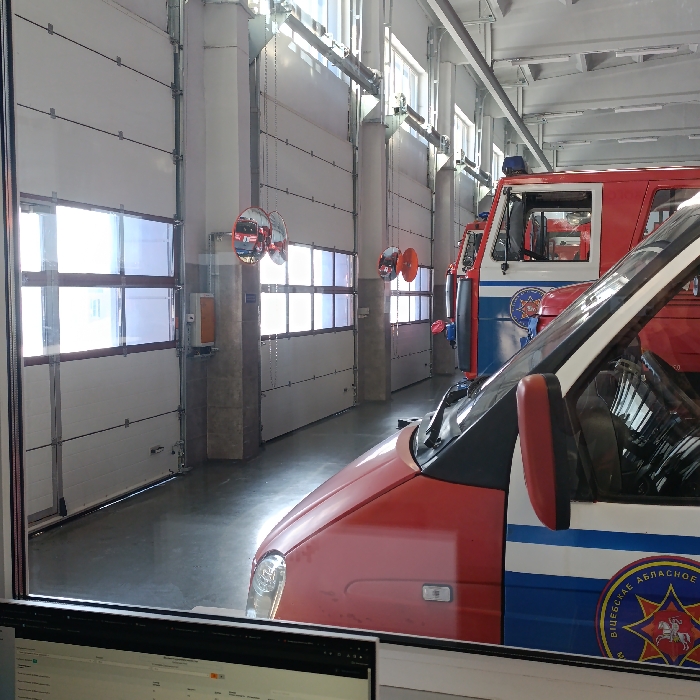 The image displays the interior of an emergency vehicle bay in Hlybokaye, Belarus, viewed through a glass partition with a computer monitor partially visible in the immediate foreground.
Two emergency vehicles are parked inside the bay. In the midground, a red and blue van, identified as a GAZelle-type, features the text "ВIЦЕБСКАЕ АБЛАСНОЕ" (Viciebsk Oblast) and a star emblem with a coat of arms on its side. Behind it, a larger red and white/blue emergency vehicle, likely a fire truck, is present with a blue light bar mounted on its roof.
The bay's left wall consists of multiple white, sectional overhead garage doors, each equipped with horizontal window panels. Structural columns separate these doors, bearing mounted convex safety mirrors. Red safety cones are suspended from chains in front of some doors. The floor is a reflective, light-colored polished concrete. Overhead, exposed ceiling structures, fluorescent lighting fixtures, and high windows providing natural light are visible. No individuals or active events are depicted within the scene.