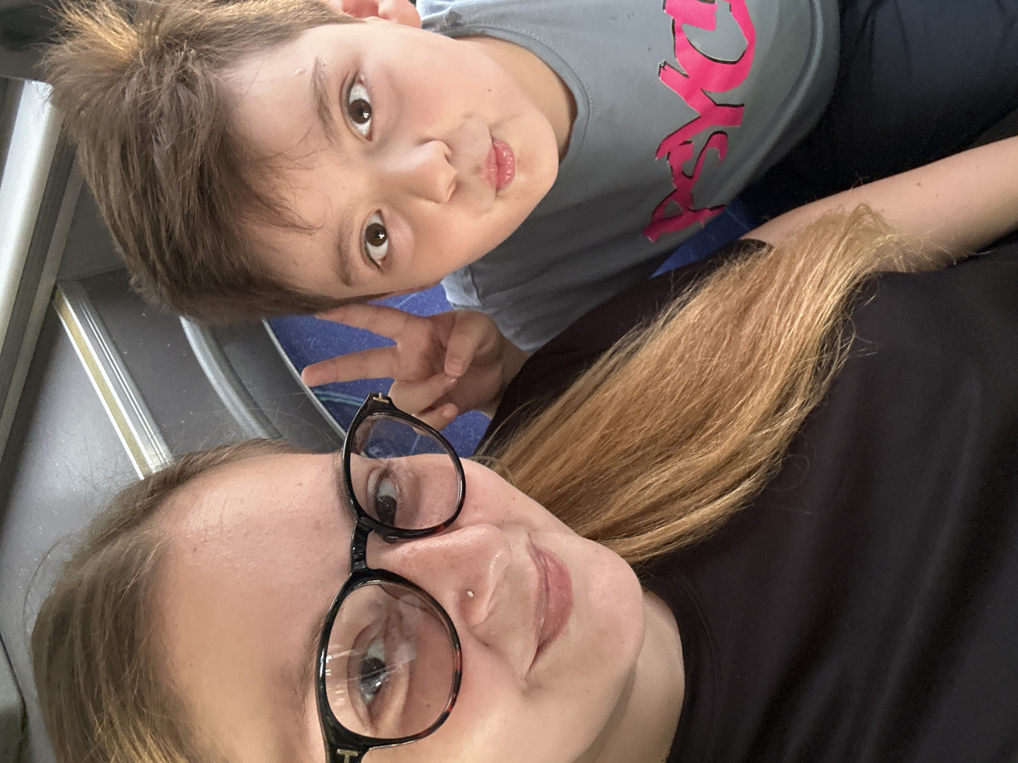 A close-up selfie shows a woman and a young boy. The woman, on the left, wears black-rimmed glasses and a black t-shirt. She has light brown hair and a nose stud. The boy, on the right, makes a peace sign with his fingers. He has short brown hair and wears a light blue t-shirt with pink lettering. The background appears to be the interior of a bus or train, with dark blue seats visible.