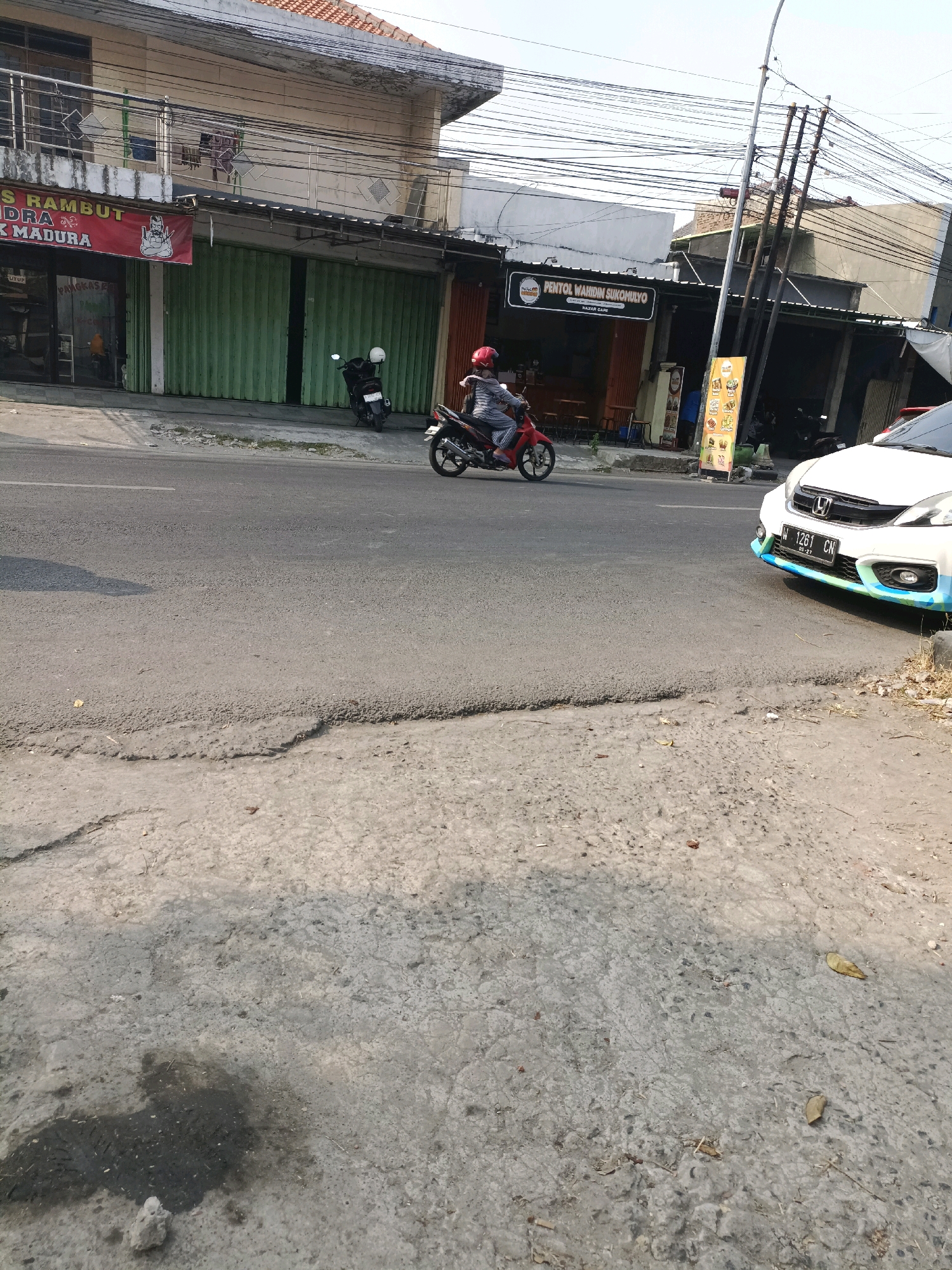 A street scene in Sukorejo, Indonesia, features a white car with license plate W 1261 CN parked on the right side of the road, facing left. To the left of the car, a person on a red motorcycle, wearing a red helmet and a gray and white outfit, is riding past. Behind the motorcycle, a white scooter is parked. In the background, buildings line the street, one with a green rolling door and a red sign that reads "S RAMBUT". Another building has a black sign that says "PENTOL WAHIDIN SUKOMULYO". A utility pole with numerous wires stands tall. The road surface is asphalt, with a rough concrete curb on the left. The overall lighting suggests it is daytime, with sunlight casting shadows.