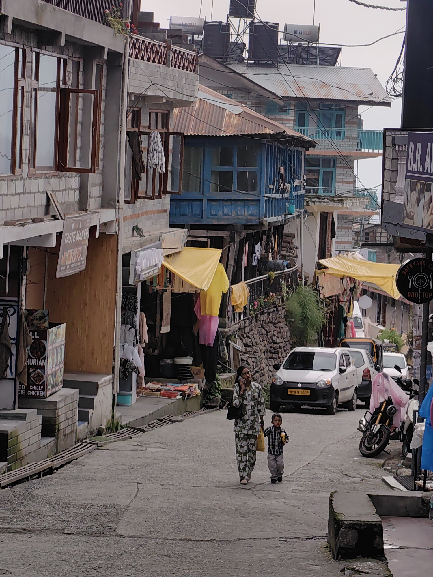 In Vashisht, India, a narrow, sloped street is lined with multi-story buildings exhibiting a mix of brick and stone construction, with many featuring corrugated metal roofs. The architecture is varied, with some structures having balconies and blue-painted window frames. Shops and businesses are visible along the street, with signage such as "TASTE MAKER" and what appears to be a restaurant menu board with "CHILLY CHICKEN" text. Yellow tarpaulins are draped over some of the storefronts, possibly for shade or protection.
Two people are walking uphill on the paved street. A woman in a patterned outfit is on her phone, holding a yellow bag in her other hand. A young boy, dressed in a checkered shirt and gray pants, walks beside her, holding a yellow object. Several parked vehicles, including cars and a motorcycle, are on the right side of the street. Laundry is hung on lines outside some of the buildings. Power lines are visible crisscrossing above the street, and water tanks are mounted on some rooftops. The overall impression is that of a busy, lived-in urban environment in a hilly region.