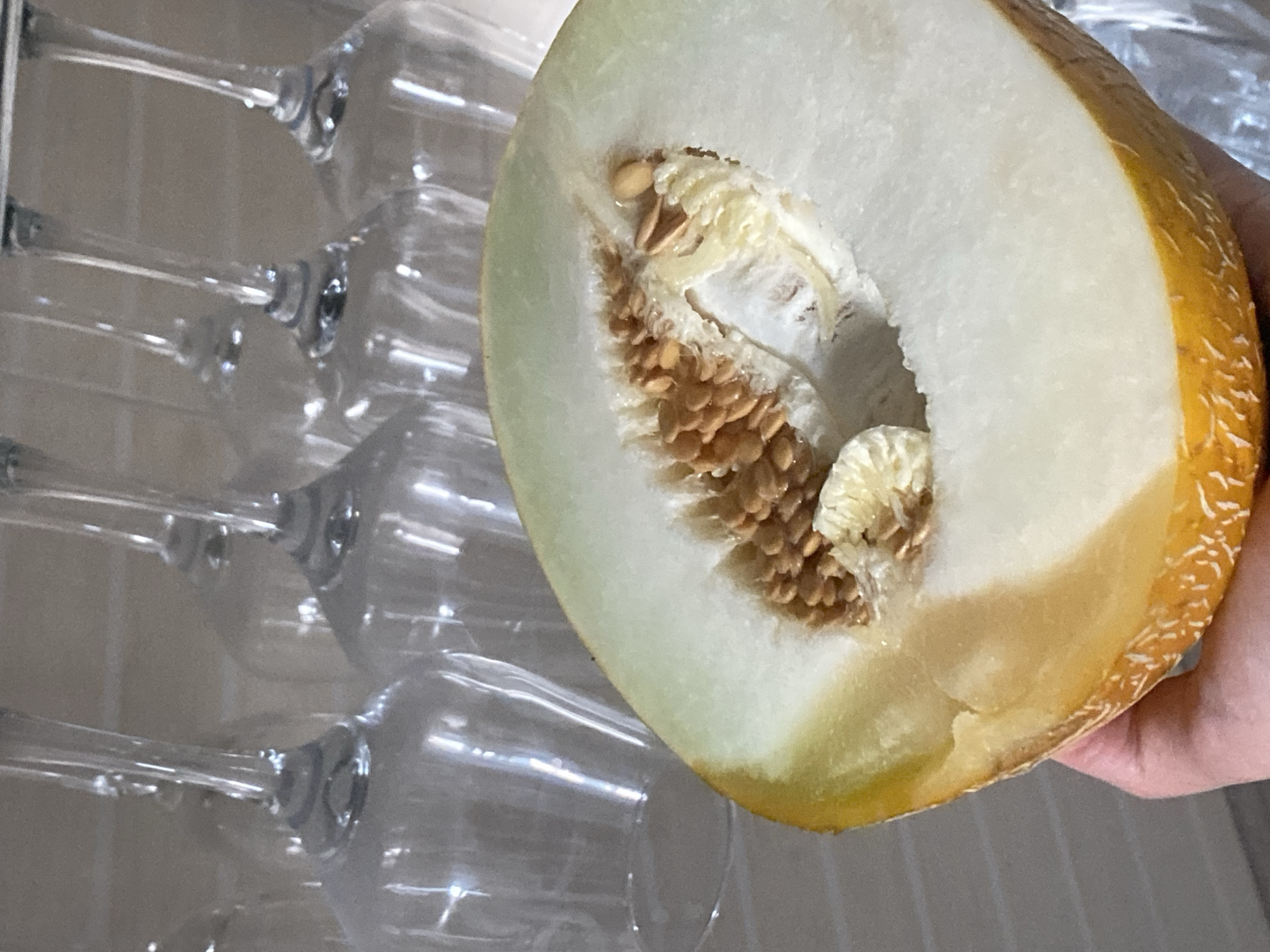 A close-up shot shows a hand holding a halved cantaloupe. The melon's interior, with its seeds and white pulp, is prominently displayed. In the blurred background, several clear wine glasses hang upside down from a rack, suggesting a kitchen or dining area. The image does not provide any information about the city of Rybnitsa, Moldova.