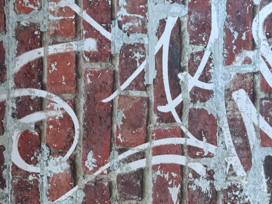 The scene depicts a weathered brick wall in Oberhausbergen, France, entirely covered with white spray paint graffiti. The wall is constructed from rectangular red bricks, showing variations in hue from dark reddish-brown to lighter orange-red. The mortar lines between the bricks are light gray and exhibit signs of cracking and flaking. The white spray paint is applied in abstract, non-figurative lines and curvilinear shapes, appearing as tags or stylized lettering that is not clearly legible. The paint application varies in density, with some areas showing solid white coverage while others are more diffuse or thin. No individuals, specific objects beyond the wall and paint, or interactions are present in the image. The environment is limited to this section of the brick wall.