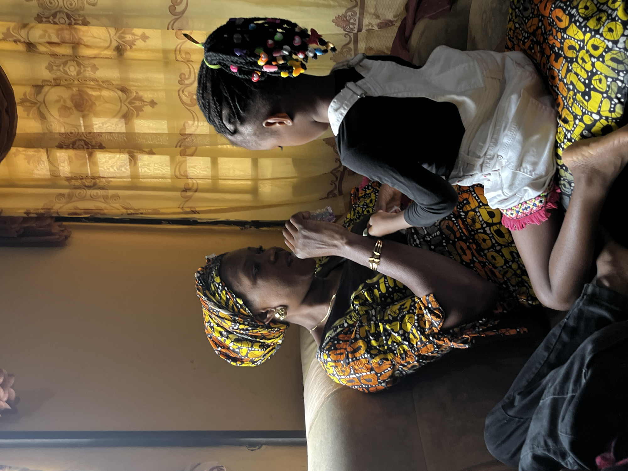 A woman and a child are indoors on a sofa. The woman wears a patterned headwrap and dress, gold jewelry, and appears to be looking at something in her hands. The child has braided hair with colorful beads and wears a black shirt and white overalls with fringe trim. The background includes patterned curtains and a wall. There is no information to infer that this photo was taken in the city of Madalla, Nigeria.