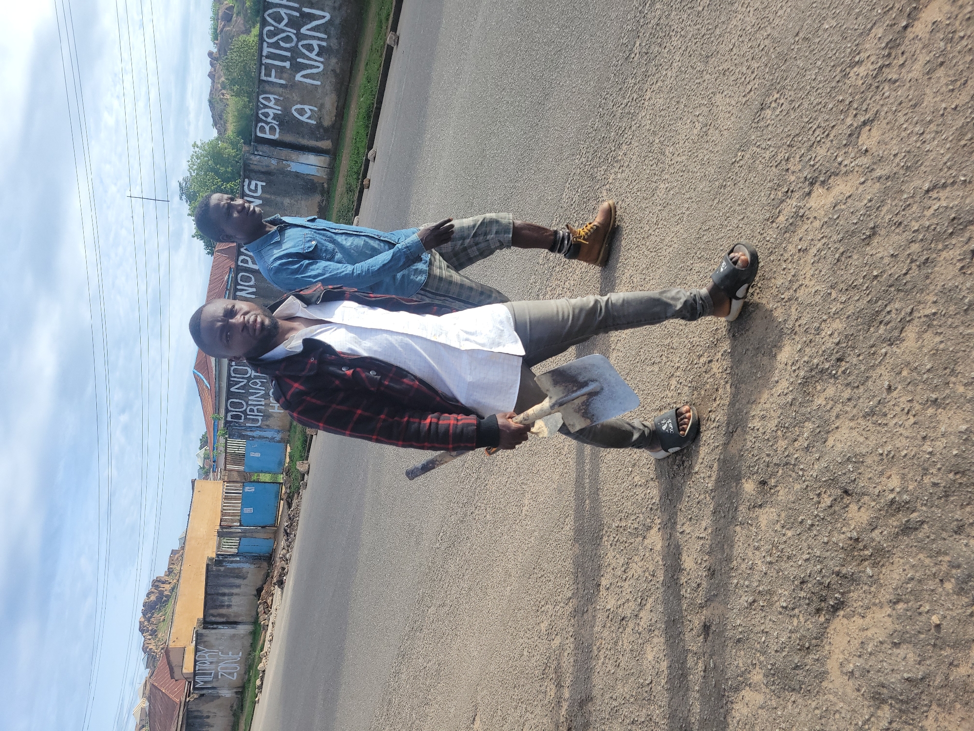 Two men walk on an asphalt road in Jos, Nigeria, under a partly cloudy sky. The man in the foreground, of dark complexion with a beard, wears a white shirt, a red and black plaid jacket, dark trousers, and black slide sandals. He holds a short-handled shovel in his right hand and looks directly towards the viewer. Behind him and to his right, a second man, also of dark complexion, wears a blue denim shirt, plaid shorts, brown work boots, and patterned socks; he looks towards his right.
On the left side of the road, a concrete wall is spray-painted with "MILITARY ZONE." Behind it, a yellow-ochre building and blue metal gates are visible, with more residential structures and a rocky hill in the distant background. On the right side of the road, a concrete wall features multiple white spray-painted inscriptions: "DO NOT URINAT" (partially obscured), "NO P" (partially obscured), and clearly legible "BAA FITSARI A NAN" (Hausa for "DO NOT URINATE HERE"). Power lines traverse the upper portion of the sky. The road surface exhibits signs of wear and scattered debris.