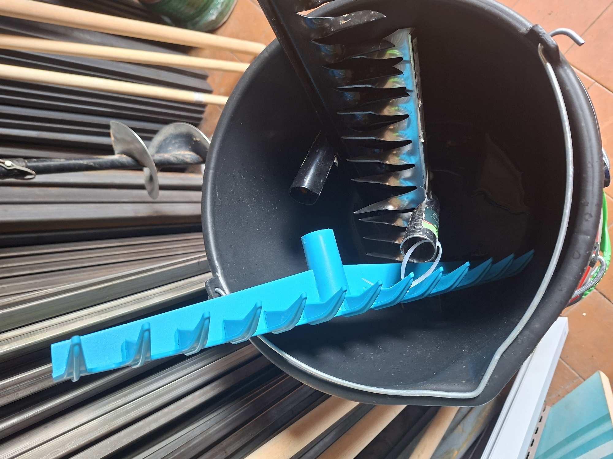 A black plastic bucket sits on a tiled floor, containing various gardening tools. A bright blue rake with a handle is positioned vertically inside the bucket, its tines facing downwards. A black metal rake with a handle is also placed within the bucket, its tines overlapping with the blue rake. Behind the bucket, an assortment of metal rods and wooden handles from other tools are visible, suggesting a storage area for gardening equipment. The overall environment appears to be an outdoor or workshop setting, possibly in Шклоў, Belarus, given the context. No people are present in the image, and no actions or interactions are depicted.