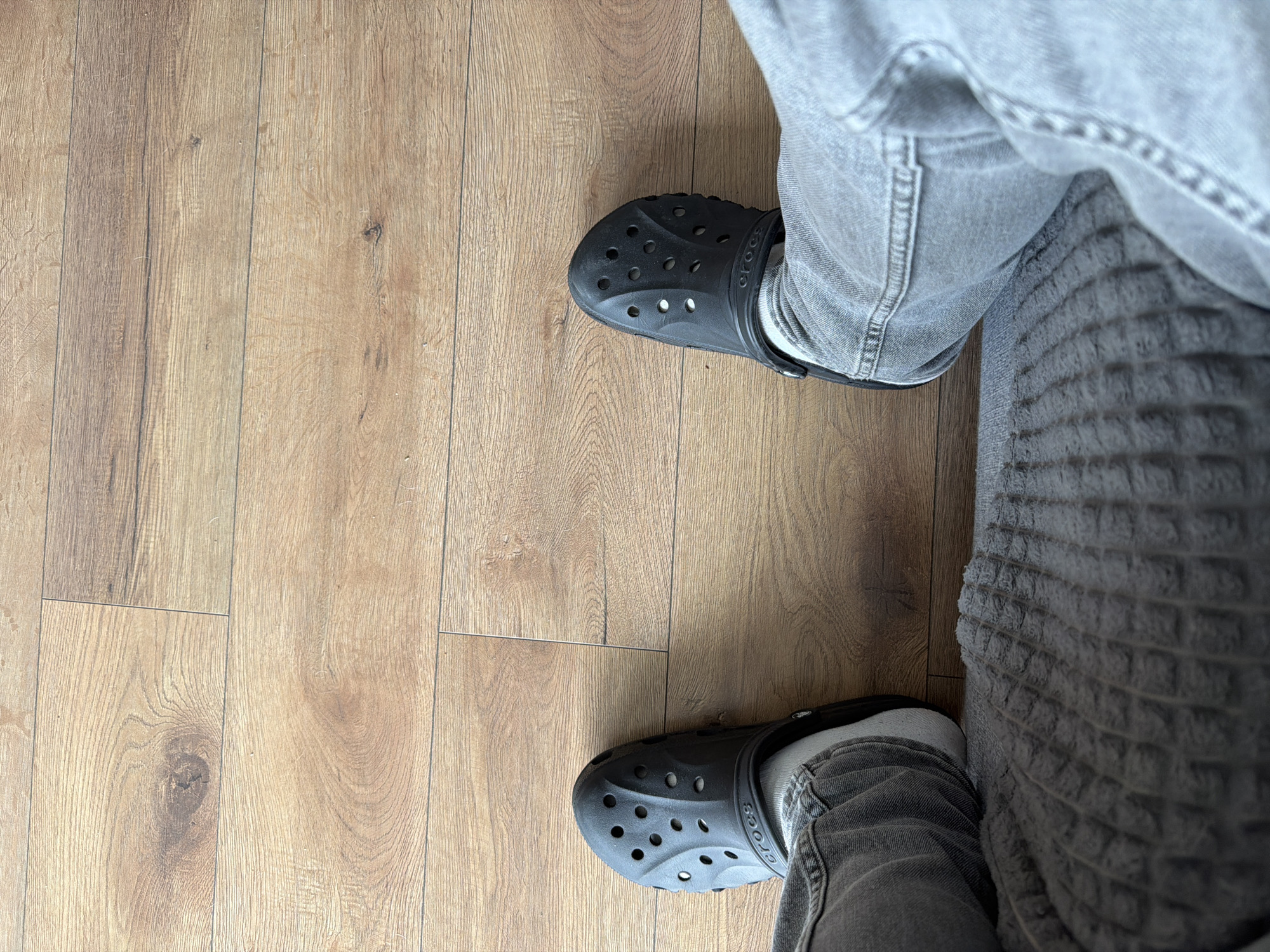 The image shows a person's legs wearing black Crocs and grey jeans, standing on a wooden floor. The perspective is from above, looking down at the feet and the floor. The floor appears to be made of laminate or wood planks with a light brown, natural wood grain pattern. The Crocs are the classic clog style with ventilation holes on the front. The jeans are a standard denim fabric, also in a grey wash. The person's left leg is bent, with the Croc on that foot pointing slightly inward. The right leg is more extended, with the Croc on that foot positioned in line with the leg. To the right of the legs, a textured grey surface is visible, possibly a blanket or upholstery. There is no clear indication of time of day, weather, or specific location beyond an indoor setting. No emotions are conveyed. The word "CROCS" is visible on the side of both shoes.