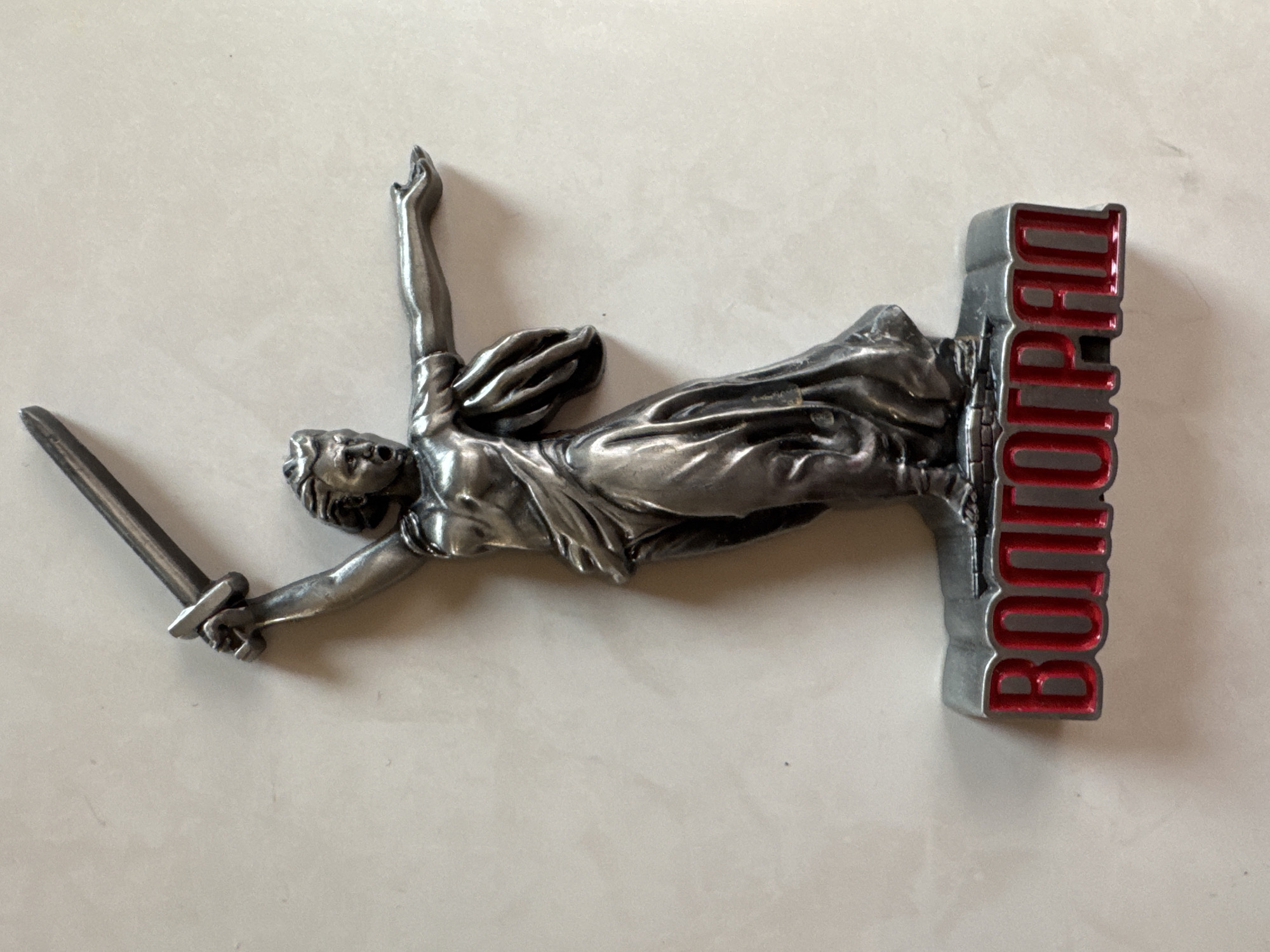 The image displays a metallic figurine of a woman holding a sword aloft, standing on a base with the word "ВОЛГОГРАД" (Volgograd) in red Cyrillic letters. The figurine is silver-toned and depicts the woman with her mouth open, appearing to shout. She is wearing a draped garment and her right arm is extended outwards. The base is grey and appears to be a raised platform. The background is a plain, light-colored surface, possibly a table or countertop. The figurine is centrally placed and slightly angled.