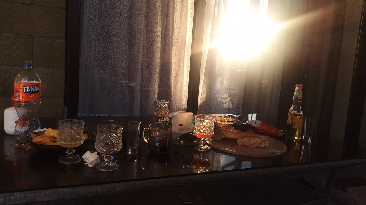 The image depicts a table set for a meal or gathering, with food and drinks. On the left is a bottle of Fanta orange soda, a bowl of chips, and several glasses. In the center, there are more glasses, one filled with a dark liquid, and a mug. To the right, there is a wooden board with bread and some snacks, and a bottle of beer. The background shows a curtain and sunlight streaming in, suggesting an indoor or covered outdoor setting. The table appears to be made of glass or a reflective material. There are no people visible in the image. The scene is set on a table, possibly outdoors on a patio or balcony, given the brick wall on the left and the view of curtains that may be covering a window or door. The lighting suggests it is either early morning or late afternoon. The location is specified as Choktal, Kyrgyzstan.