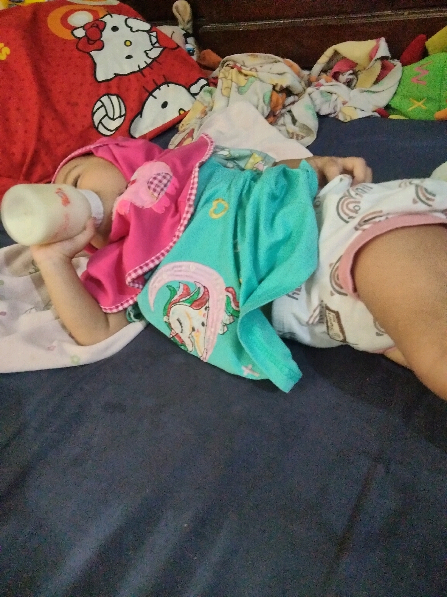 A young child wearing a pink hood and a turquoise onesie with a unicorn graphic is lying on their back and drinking from a baby bottle. The child is on a dark blue surface, possibly a bed or floor, and is surrounded by blankets and stuffed animals. The blanket closest to the child has a Hello Kitty graphic. The overall scene appears to be indoors. The city of Suka Sari, Indonesia is not depicted in this image.