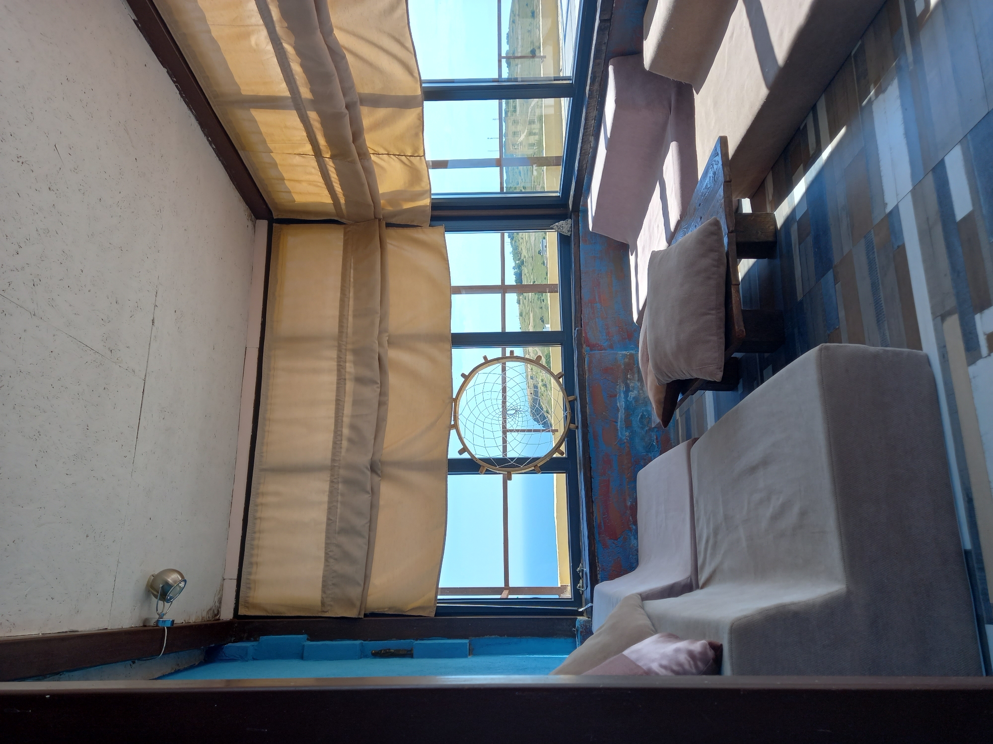A sunlit indoor room features a U-shaped seating area with beige cushions, a dark wood coffee table with a brown pillow, and a patterned tiled floor. Large windows offer a view of the sea and a hilly landscape. The walls are painted a vibrant blue with some visible texture, and the ceiling has a textured white surface with a small spotlight. A circular dreamcatcher-like ornament hangs in one of the windows. The room appears to be a lounge or seating area with an oceanic view.