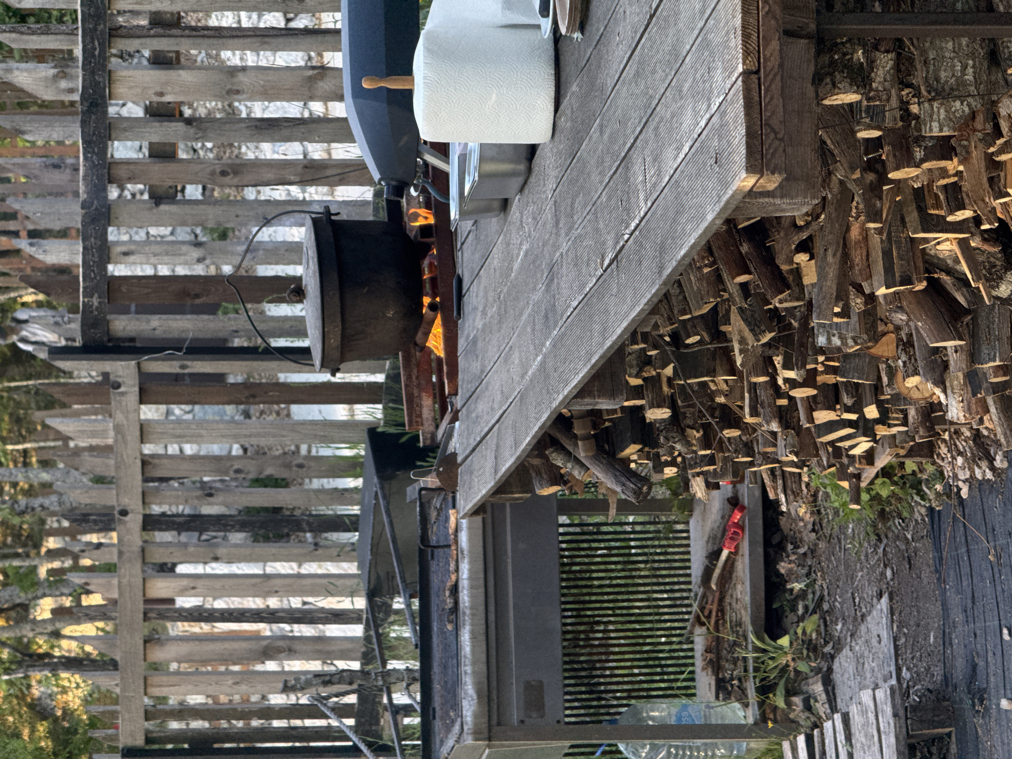 The image depicts a weathered outdoor setting in Bukovik, Montenegro. A wooden fence stands to the left. A grill with a smoker attached is central. A roll of paper towels sits on a wooden table next to the grill. Wood is piled under the table. An axe is also visible on the ground near the wood pile. Flames can be seen in the smoker.