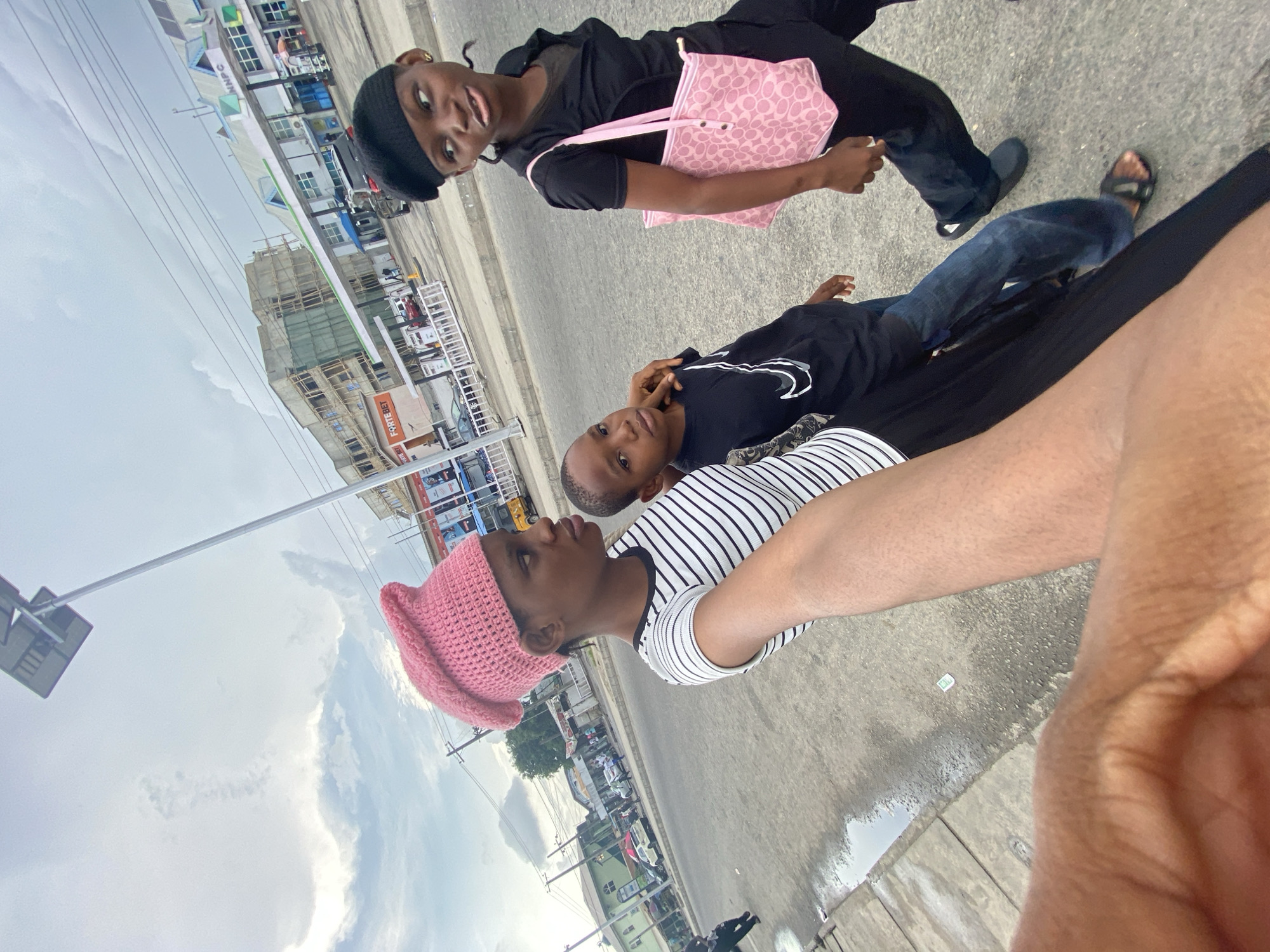 A medium shot, taken at an unusual angle, shows three people outdoors on a city street in Unknown, Unknown. The perspective is tilted so that the individuals are positioned diagonally across the frame, appearing to be lying down.
At the bottom of the frame, a person wearing a striped shirt and a pink knitted hat faces the left. Beside them, a younger person wearing a black t-shirt and jeans rests their head in their hand. Above them, a third person is seen with a black beret, black top, and jeans, carrying a pink patterned handbag.
The environment includes a paved street with buildings and street signs in the background. The sky is partly cloudy. The camera angle suggests the photo may have been taken by the person in the striped shirt, with their arm extended into the frame.