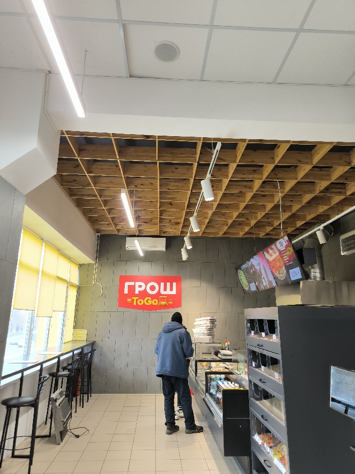 The image captures the interior of a modern fast-food or takeaway establishment, likely a cafe or convenience store with a hot food section, located in Illintsi, Ukraine. The setting features a blend of industrial and contemporary design elements.
The scene is dominated by contrasting ceiling designs: the front half of the room has a standard white acoustic tile ceiling with a recessed circular light, while the back half features an eye-catching wooden grid ceiling with square compartments, through which some lighting is visible. Various linear LED strip lights and white track lighting fixtures with adjustable spots illuminate the space.
The walls are clad in light grey concrete-like panels or blocks, giving an industrial feel. A large red sign is prominently displayed on the wall, reading "ГРОШ" (GROSH) with "=ToGo=" underneath in white lettering, indicating the establishment's name and takeaway service. Above this sign, an air conditioning unit is mounted.
Along the left side of the room, a large window allows natural light to enter, though yellow roller blinds are mostly drawn. A long, high bar table runs parallel to the window, accompanied by several black high-backed stools, suggesting a casual dining or waiting area. A small portable electric heater is visible beneath the window bar.
In the center-right of the image, a single person is seen from behind, wearing a dark beanie and a blue-grey hooded jacket over dark trousers. They are standing in front of a long, glass-fronted refrigerated display case filled with various food items. Behind this counter, stacks of white boxes, possibly pizza boxes, are visible. To the right of the display case, a multi-tiered black shelving unit holds more packaged items. Above the food preparation area, several digital menu boards display colorful images of food, such as pizzas, along with text and prices, likely in Ukrainian.
The floor is tiled with light-colored square tiles. The overall impression is a clean, brightly lit, and functional space designed for quick service. The time of day appears to be daytime, indicated by the natural light, even with the blinds down.