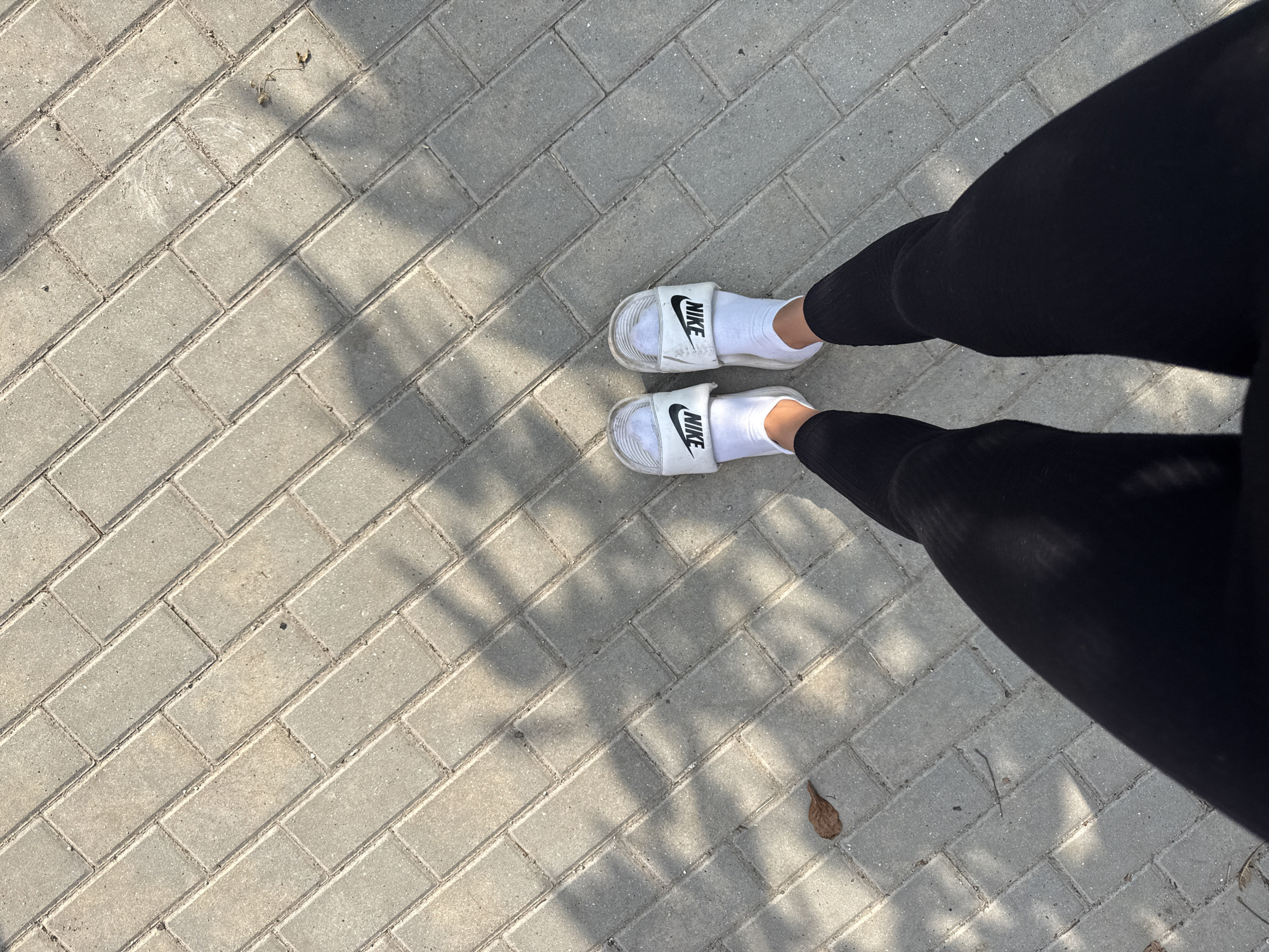 The image shows a person's legs and feet wearing black leggings, white socks, and white Nike slides, standing on a paved walkway composed of rectangular gray bricks laid in a herringbone pattern. The scene appears to be outdoors, as evidenced by the sunlight casting shadows of foliage on the ground. There is a single brown leaf on the walkway. The context suggests a casual setting in the city of Basarabeasca, Moldova.