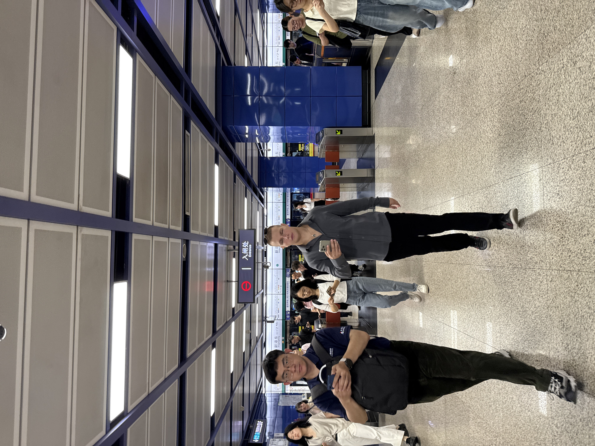 In the Haizhu District, China, an interior shot captures a bustling metro station. A group of individuals are seen walking on a polished, speckled floor beneath a modern ceiling with integrated lighting and blue structural beams. A prominent blue tiled column stands to the right. In the foreground, a man in a dark grey shirt and black pants walks towards the viewer, holding a smartphone. To his left, a man in a blue shirt and black pants, wearing glasses and earbuds, also walks forward, looking at his phone while carrying a backpack. Behind them, other commuters are visible, some also engaged with their phones. An "Entrance" sign with Chinese characters is mounted overhead. Metal turnstiles are present in the background, suggesting a paid access area.
