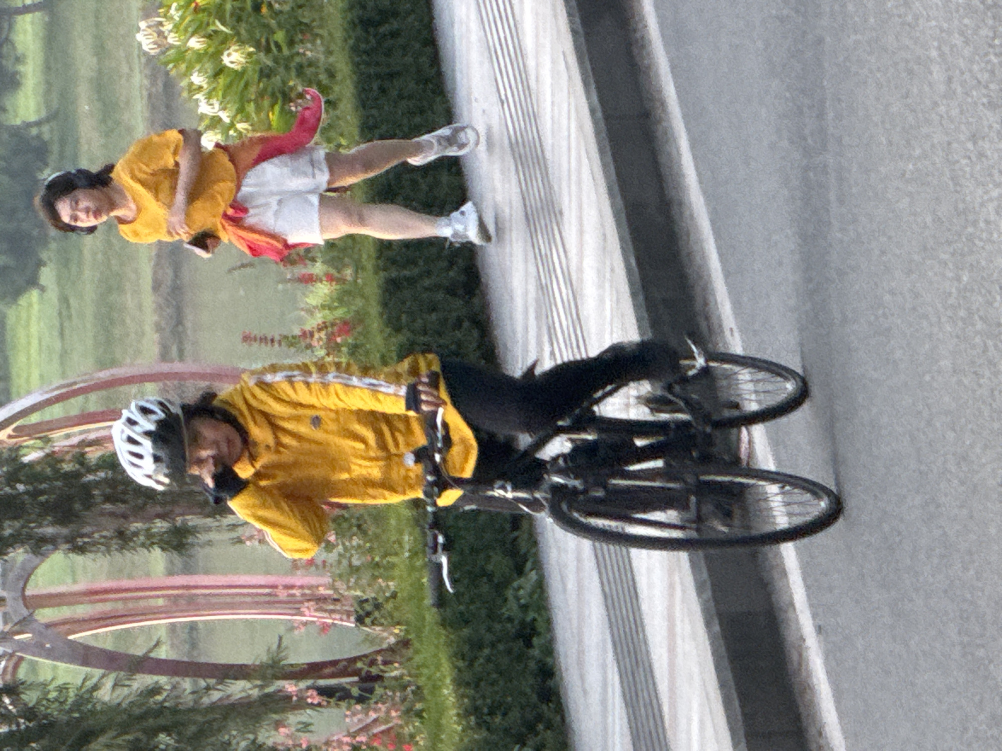 In Da Lat, Vietnam, on a paved walkway beside a body of water, a person on a bicycle wearing a yellow jacket and white helmet is captured in motion, with their right hand near their face as if wiping away sweat or an irritant. Behind them, another individual is running, wearing a yellow top and white shorts, with headphones on. The background features lush green foliage and decorative arches.