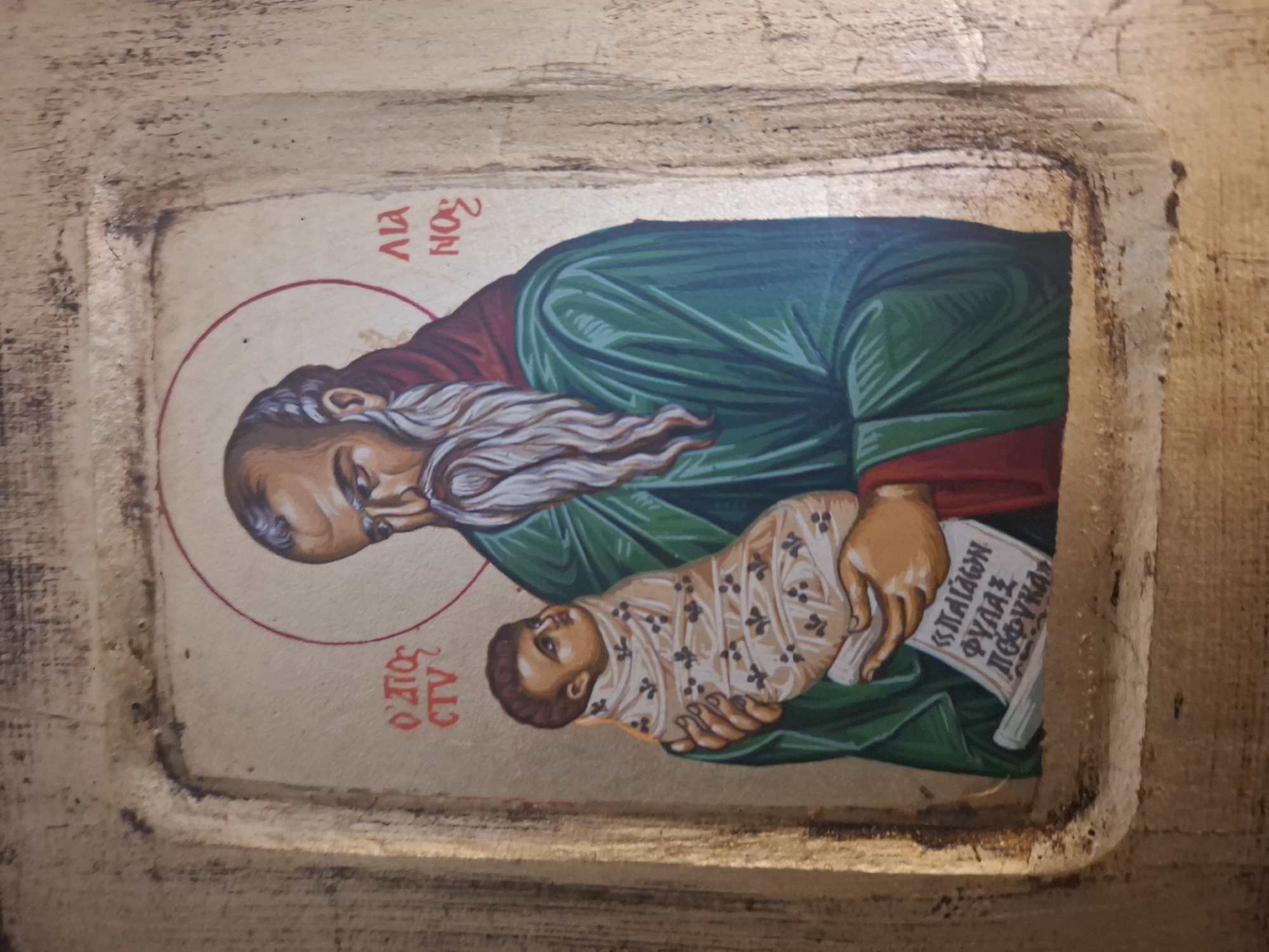 An icon painting depicts Saint Simeon holding the infant Jesus. The figures are rendered in a traditional Byzantine style with halos. The man, identified by Greek lettering as "OSIOS SI...MON" (likely Saint Symeon) on the left and "ΛΙΛΑ ΝΟC" (possibly an abbreviation or inscription) on the right, has a long grey beard and wears a red robe over a green garment. He holds a swaddled infant, presumably Jesus, who looks upwards. Below the figures, a scroll with Greek text is visible. The painting is framed by a weathered, carved wooden border. The icon is situated within a carved recess, possibly a niche or part of a larger architectural feature. The background of the icon is a muted gold. The overall appearance suggests an aged religious artifact, likely in a place of worship or private devotion. This icon is located at the Église Saint-Nicolas in Montreuil, France.