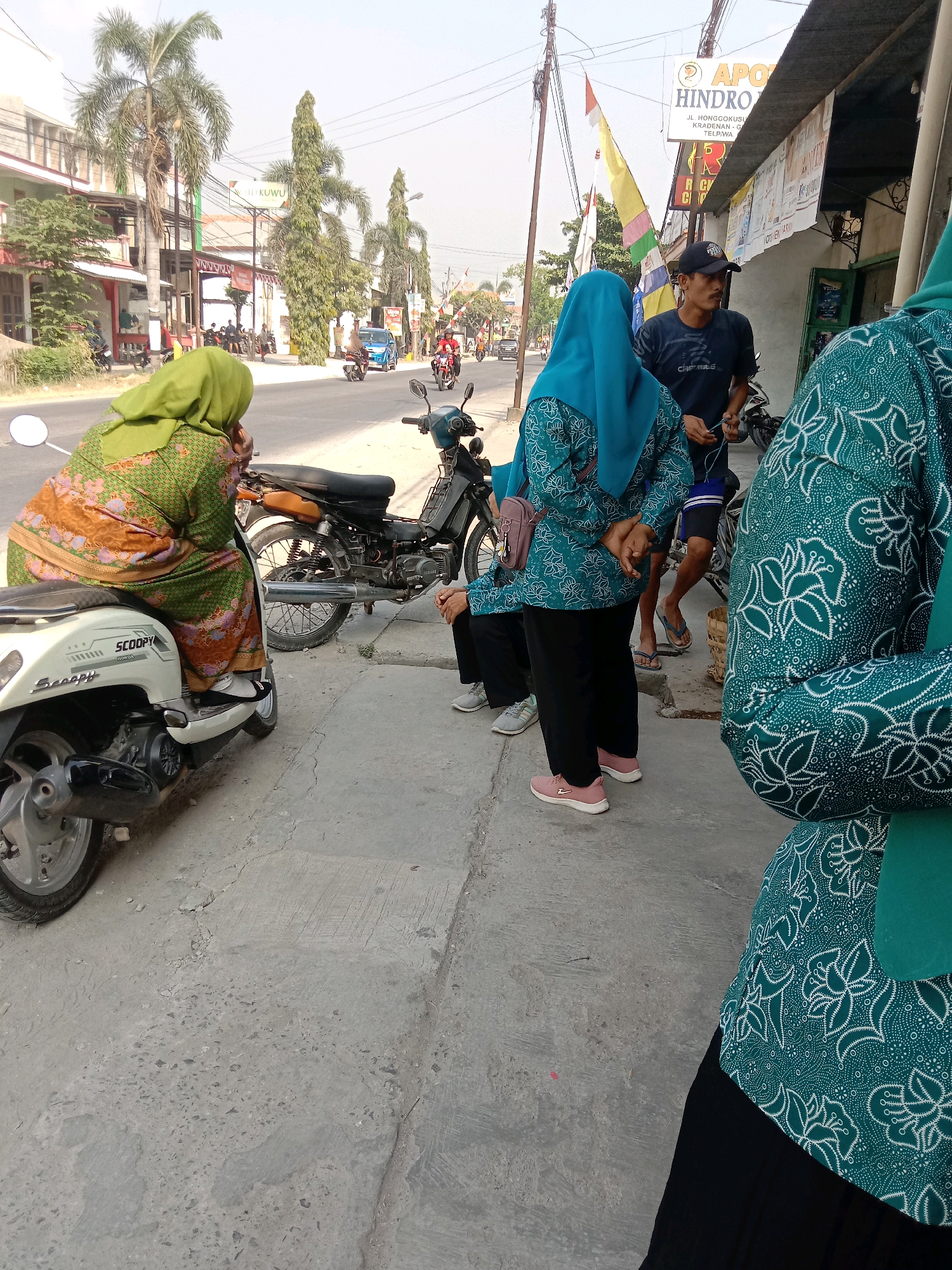 In the city of Kalisari, Indonesia, a street scene unfolds with several individuals and vehicles. A woman in a green hijab and a patterned orange and green dress sits on a white Honda Scoopy scooter parked along a concrete sidewalk. To her right, two women, both wearing teal hijabs and matching teal and white patterned tops with black pants, stand on the sidewalk. One of them is interacting with a man wearing a dark blue t-shirt and a black baseball cap, who is holding some wires. A second motorcycle, a black Yamaha, is parked behind the women. In the background, a street lined with buildings and palm trees is visible, with traffic including cars and motorcycles. A sign for "APOTIK HINDRO" is mounted on one of the buildings. The sky is clear and bright.