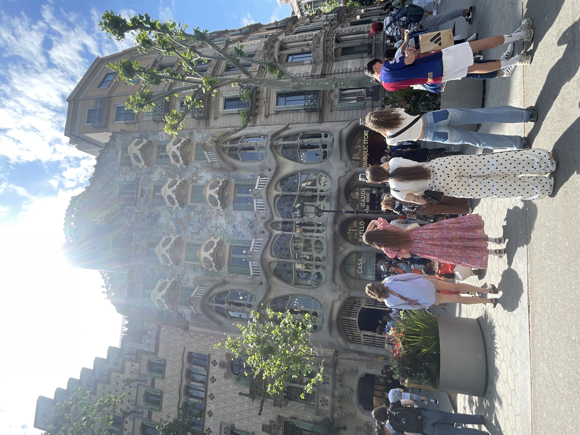 The image presents a bustling street scene in Barcelona, Spain, centered around Antoni Gaudí's iconic Casa Batlló. The building's unique facade is a prominent subject, characterized by its wavy, organic shapes, colorful mosaic-like tiling, and distinctive bone-like balconies.
Numerous people are visible on the sidewalk, appearing to be a mix of tourists and passersby. Many are standing, looking up at the architectural marvel, with several individuals actively capturing photos or videos with their smartphones. Other people are strolling, contributing to the lively atmosphere. One person is seen carrying an Adidas shopping bag.
The setting is an urban environment on a bright, sunny day, with clear skies featuring scattered clouds. The strong sunlight creates distinct shadows on the pavement and produces a noticeable lens flare effect on the left side of the image, suggesting late morning or afternoon. The overall mood is active and engaged, reflecting the building's status as a popular point of interest.
Visible text includes "SIMBOLIC STORE" and "GAUDÍ" on storefront signage. The name "CASA BATLLÓ" is also partially discernible on a plaque or sign attached to the building. An "ADIDAS" logo is visible on a shopping bag, and some text, possibly "LOTT" or "Lo," is seen on a person's socks.