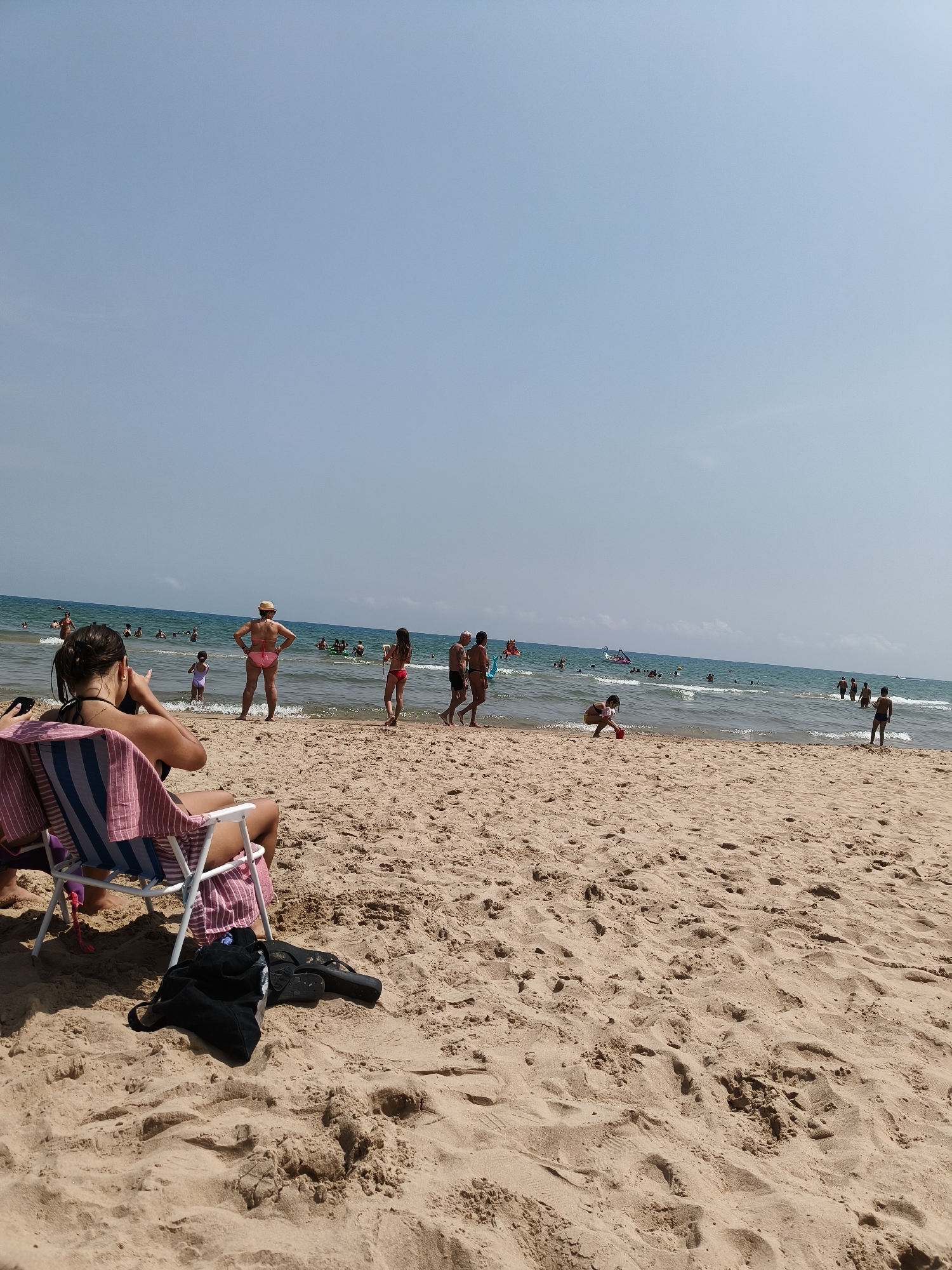 This is a wide-angle, eye-level shot of a sandy beach on a sunny day. The scene is set in Grau I Platja, Spain. In the foreground, a woman with dark hair is seated in a striped beach chair, facing away from the camera and towards the water, appearing to take a photo with her phone. Her bag and a pair of flip-flops rest on the sand next to her chair.
The beach is populated with many people. In the middle ground, several individuals are visible in the shallow water, some wading, some swimming, and a few children playing. A woman in a straw hat and pink bikini stands facing the sea, her hands on her hips. Further out, more people are scattered across the water, and a few are visible on the shoreline. The ocean stretches to the horizon under a clear, pale blue sky. The sand itself is textured with footprints and small undulations.