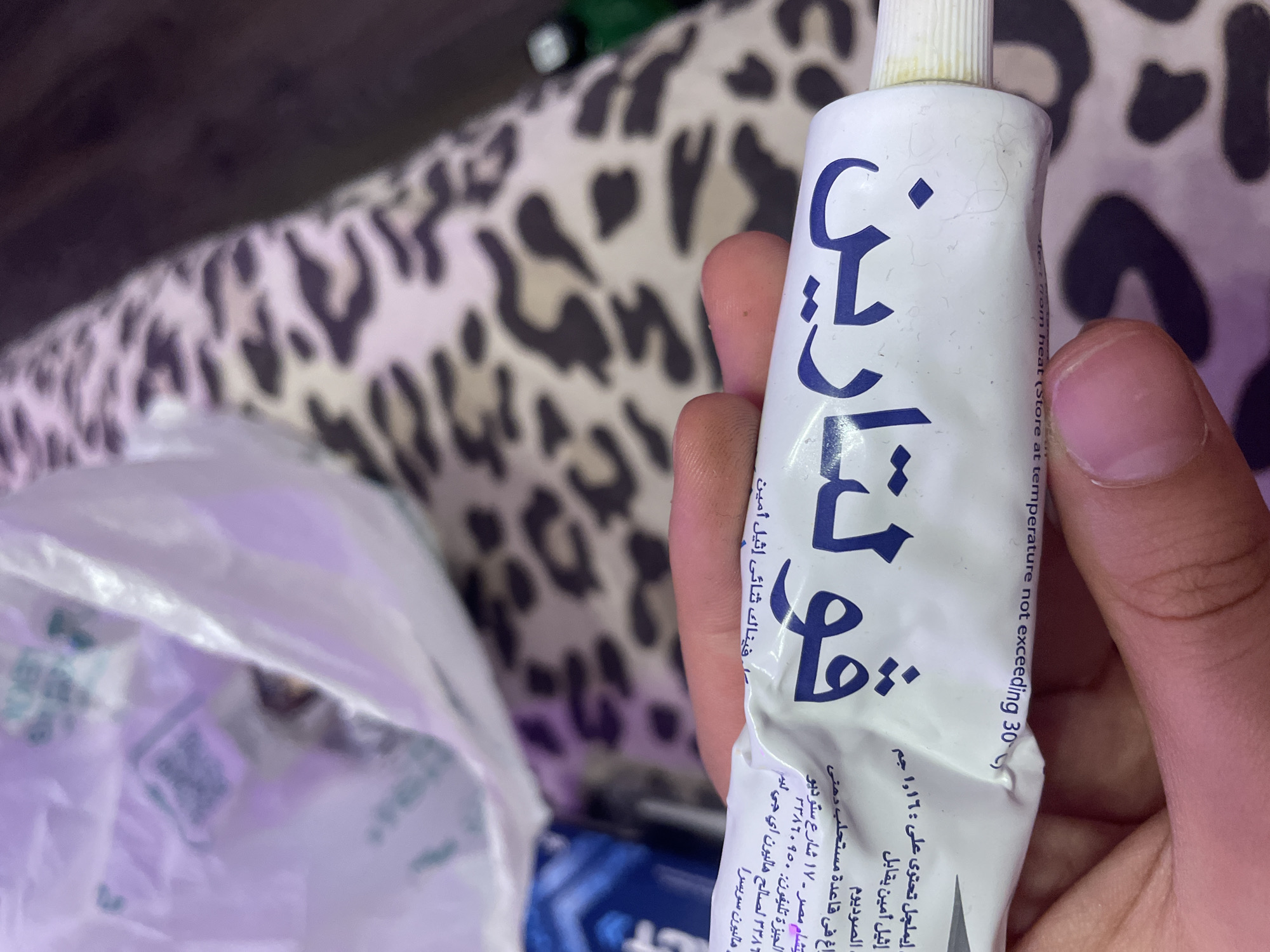 The image shows a close-up of a white tube of ointment with Arabic text and some English text. The tube is held by a person's hand, with fingers visible on the right side. The ointment tube has blue Arabic lettering that appears to be a brand name, and also some smaller text in Arabic and English. Part of a white plastic bag is visible in the background, as is a patterned fabric. The English text on the tube includes "Store not exceeding 30 C". There is no information or visual evidence in the image that indicates the location is Vaganovo, Russia.