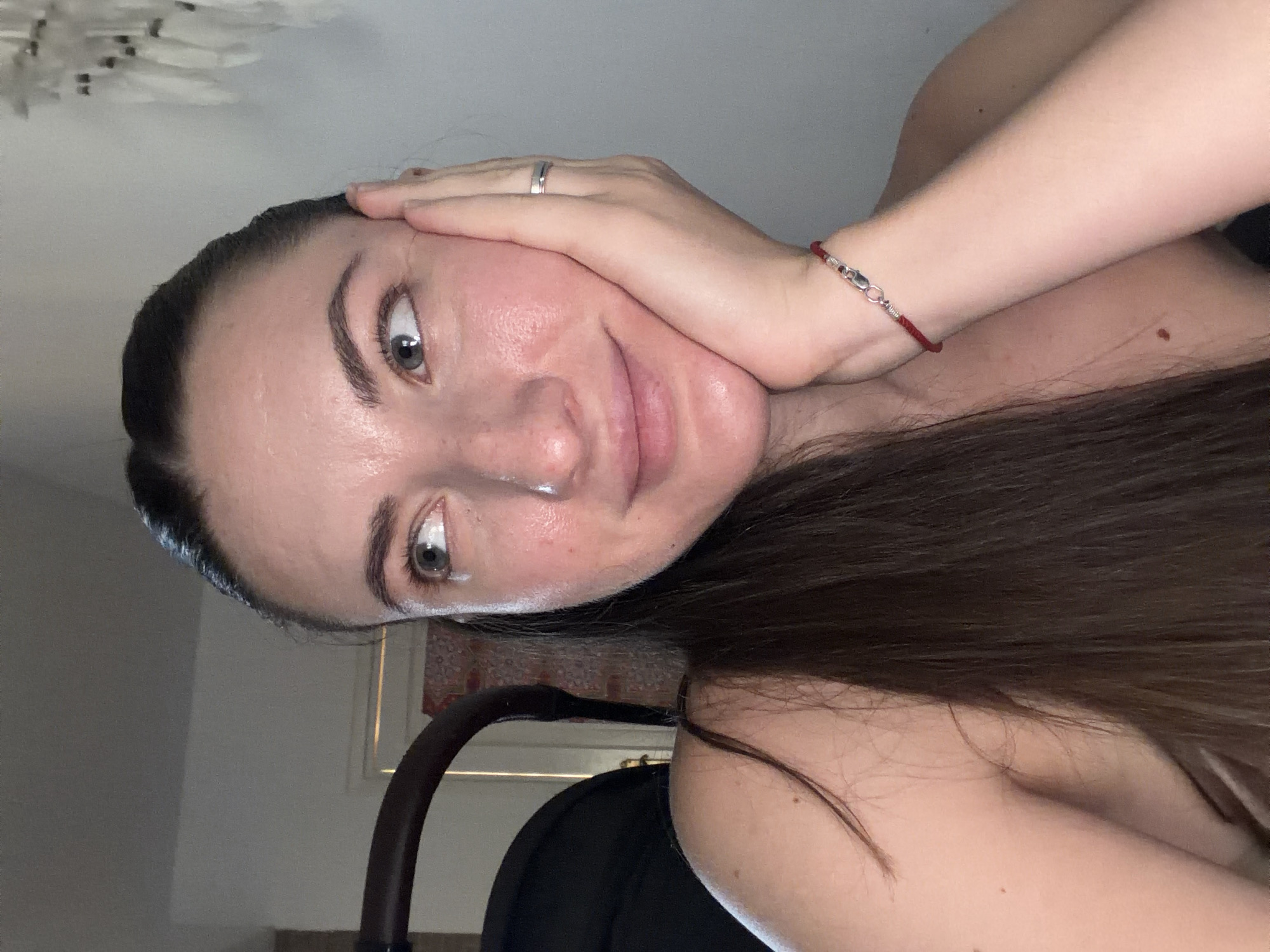 A woman with her right hand resting on her cheek is shown in a medium close-up shot. Her head is turned to the left, and her eyes are looking in the same direction. She has long, dark brown hair. She is wearing a simple silver band on her ring finger and a red string bracelet with a silver clasp on her left wrist. She is also wearing a thin necklace. The background is out of focus and appears to be an indoor setting. There is a dark object to the left of the woman that resembles a stroller. The overall lighting suggests it is indoors, with a light source coming from the right side of the frame. The image was taken in Sharm El Sheikh, Egypt.