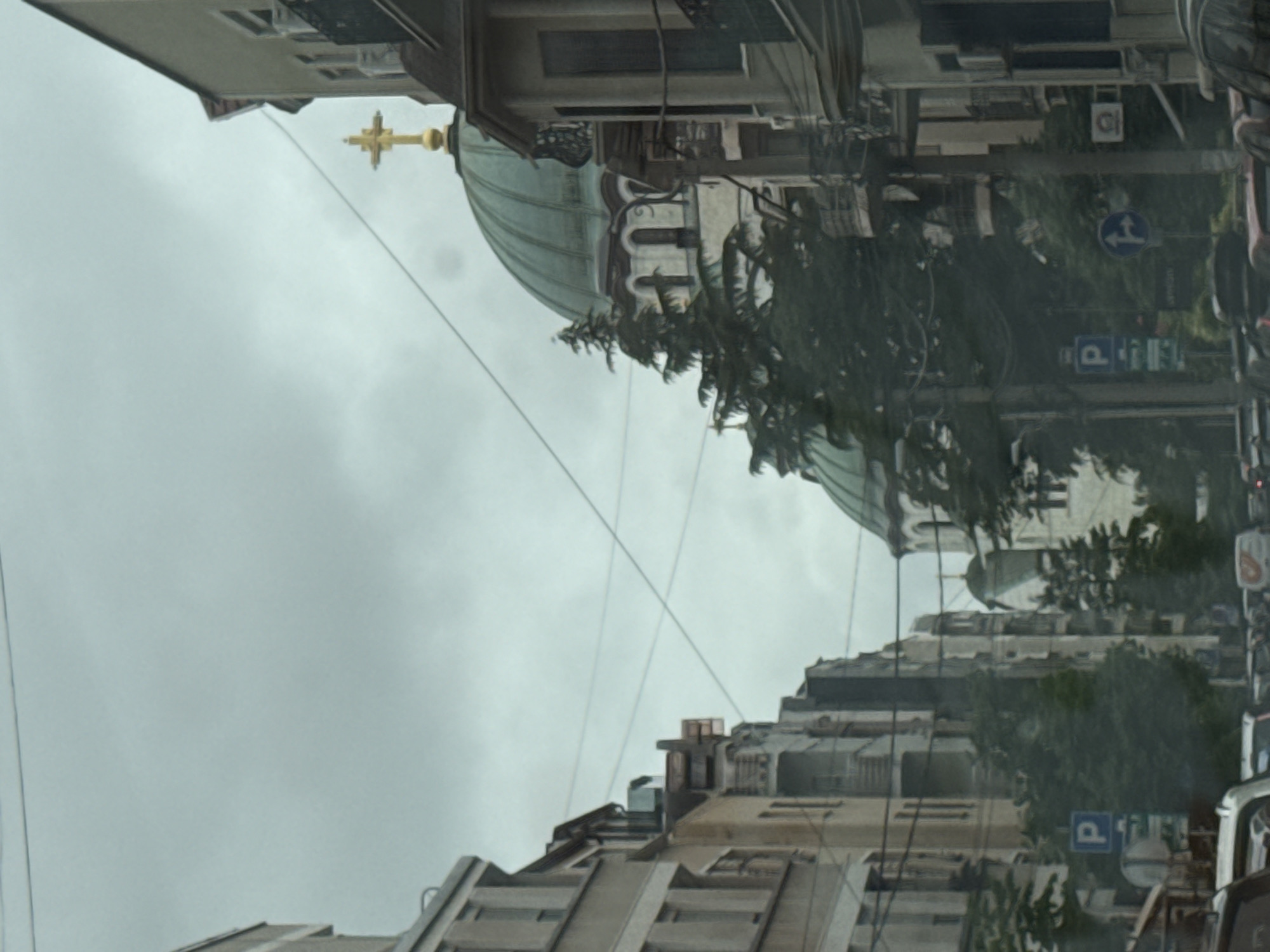 The image depicts a view down a city street with buildings on either side and a large domed church in the background. The sky is overcast and gray. The buildings appear to be residential or commercial, with multiple stories and windows. There are trees lining the street and some power lines are visible overhead. In the foreground, several cars are present, suggesting that the photo was taken from a moving vehicle. There are also street signs visible, including one with a blue "P" indicating parking. The architecture of the domed church, particularly its green dome and golden cross, is distinctive and characteristic of Orthodox Christian churches. The overall impression is of an urban environment with a significant religious structure at its center. The scene is consistent with Vracar Urban Municipality in Serbia, known for its churches and residential buildings.