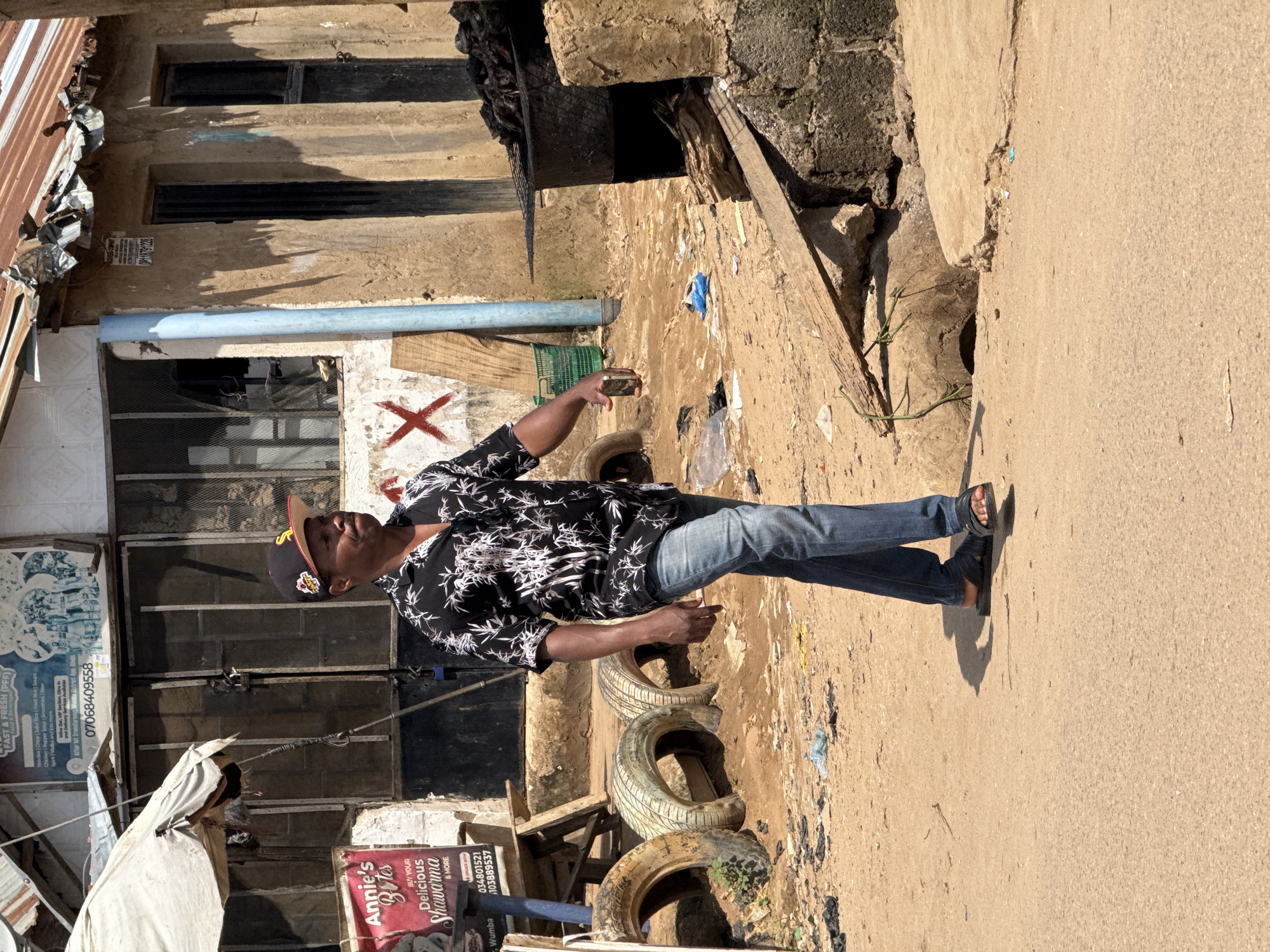 A man is walking on a dusty street in Abuja, Nigeria. He is wearing a black shirt with a white floral pattern, blue jeans, and black sandals. He has a black baseball cap on and is holding a smartphone in his right hand. In the background, there are several buildings with corrugated metal roofs, some of which appear to be stalls or shops. There are also some tires, wood planks, and debris scattered on the ground. A grill with food is visible to the right of the man. The scene depicts a typical street view in a bustling urban environment.