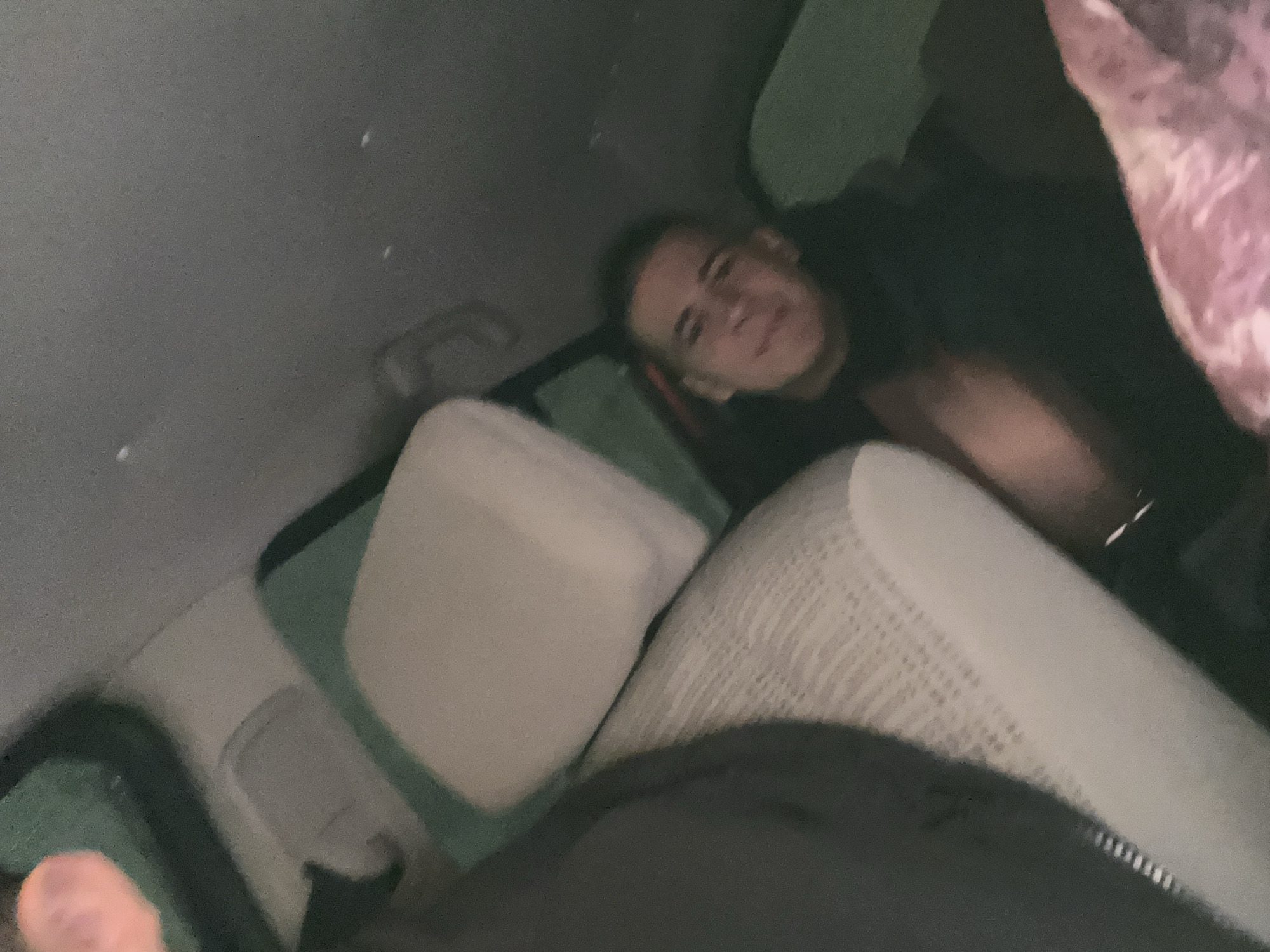 The scene depicts the interior of a vehicle, likely a car or van, under low light conditions. One male individual, appearing to be a young adult, is visible in the middle-right of the frame. He is wearing a dark-colored upper garment and is looking towards the camera with a slight smile. A partially visible, out-of-focus, orange/pink object, possibly a hand or arm, is present in the top-left corner. The vehicle's interior features include a grey headliner, a grab handle near the ceiling, a light-colored, textured seat dominating the left foreground, and a light-colored cushion or pillow with a green/teal covering behind it. A pink and white patterned fabric object is partially visible in the bottom-right. The specific location within the city of Unknown, Unknown, cannot be determined from the image.