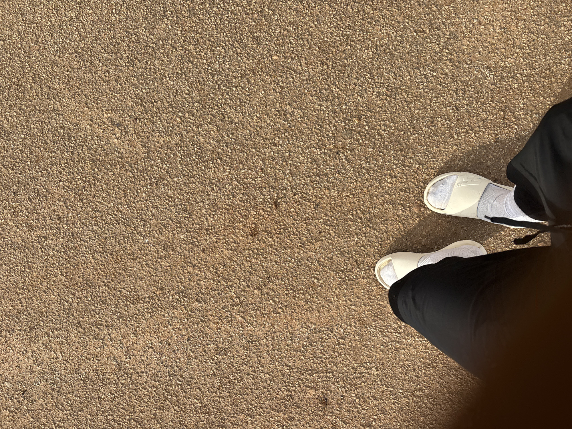 The image shows a person's feet and lower legs standing on a paved surface. The paving appears to be made of small gravel or pebbles set in a binding agent. The person is wearing white socks and light-colored sandals. Black pants cover the legs. The ground is a brownish-beige color. There is no information in the image to suggest the location is Bukuru, Nigeria. The environment appears to be outdoors.