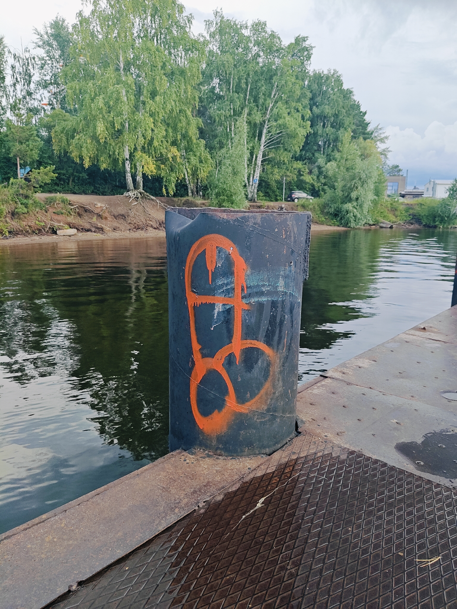 A cylindrical, dark-colored bollard is positioned on the edge of a metallic platform. The bollard has orange graffiti depicting a phallic symbol sprayed on its surface. The platform is next to a body of water, which reflects the green foliage of trees lining the opposite bank. The environment suggests a waterfront area, likely in a semi-urban or natural setting, with a pathway or embankment visible behind the trees. The sky is overcast with visible clouds. Based on the context provided about Chaikovsky, Russia, this scene is likely situated along the Kama River or a similar waterway within or near the city.